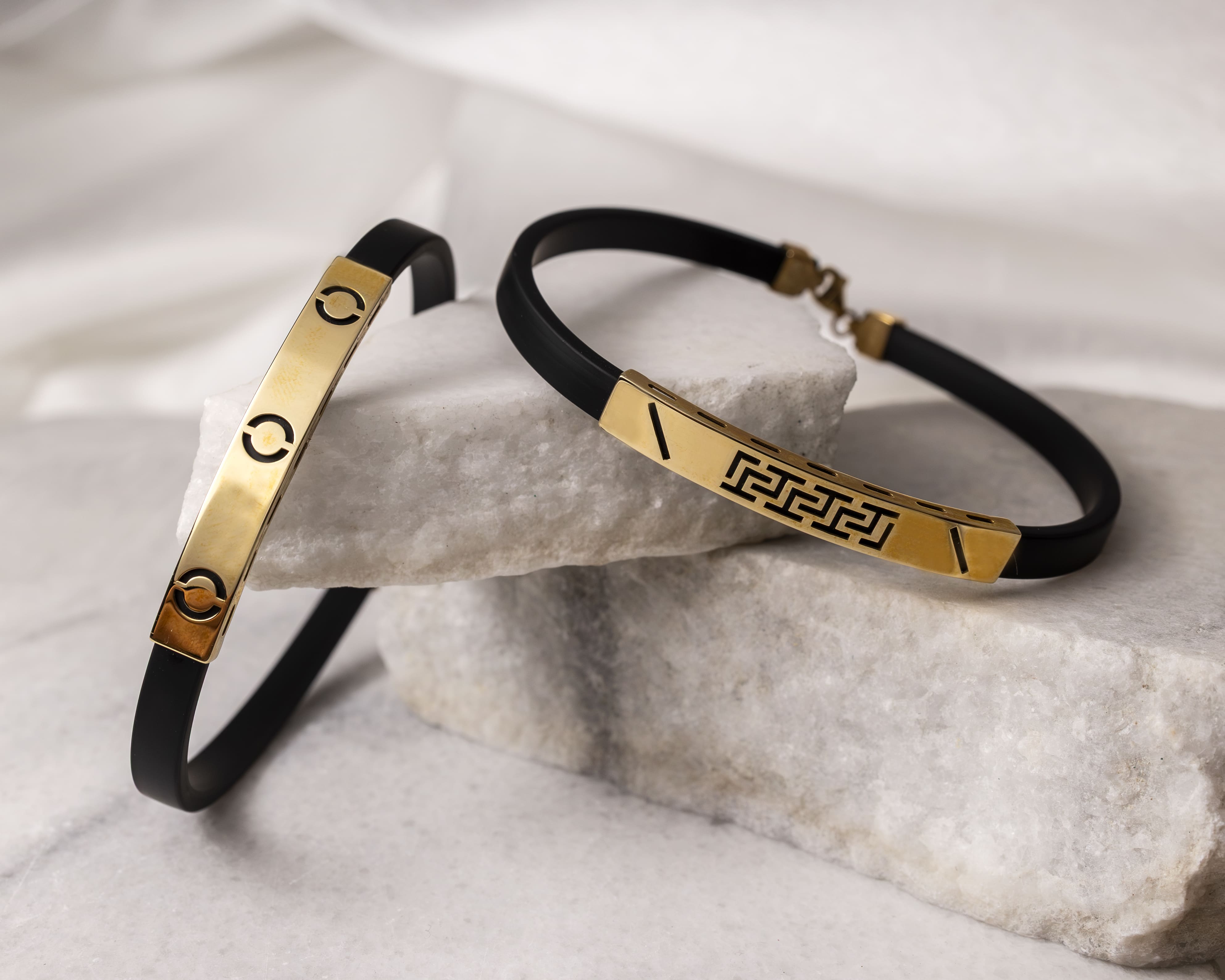 Two black bracelets with gold decorative plates placed on white stone slabs.