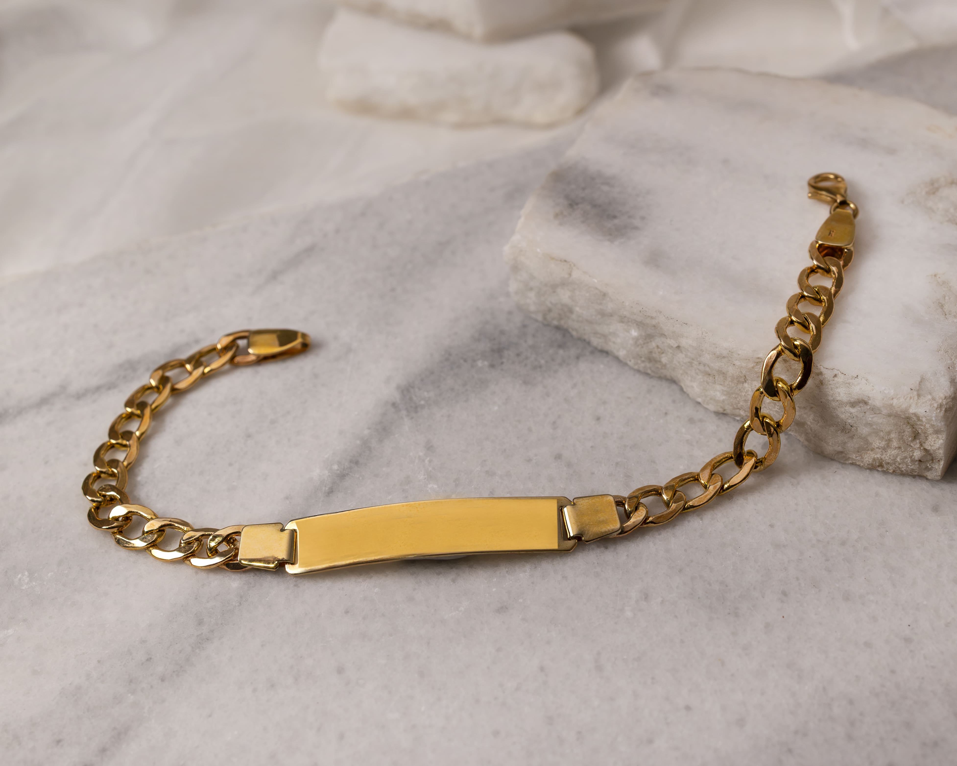 Gold chain bracelet with a blank rectangular plate on a white marble surface.
