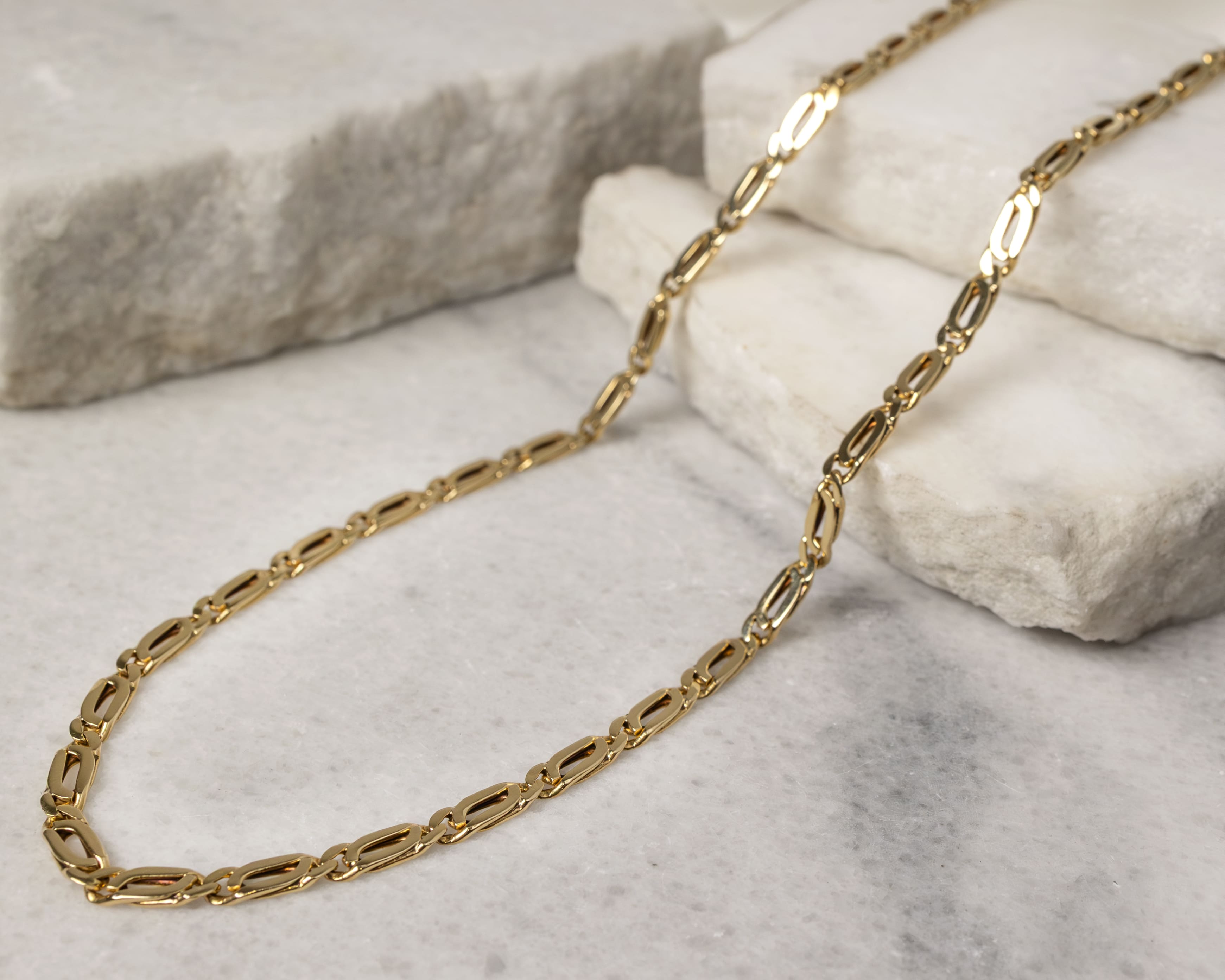 Gold chain necklace with elongated oval links displayed on white textured stone surfaces.