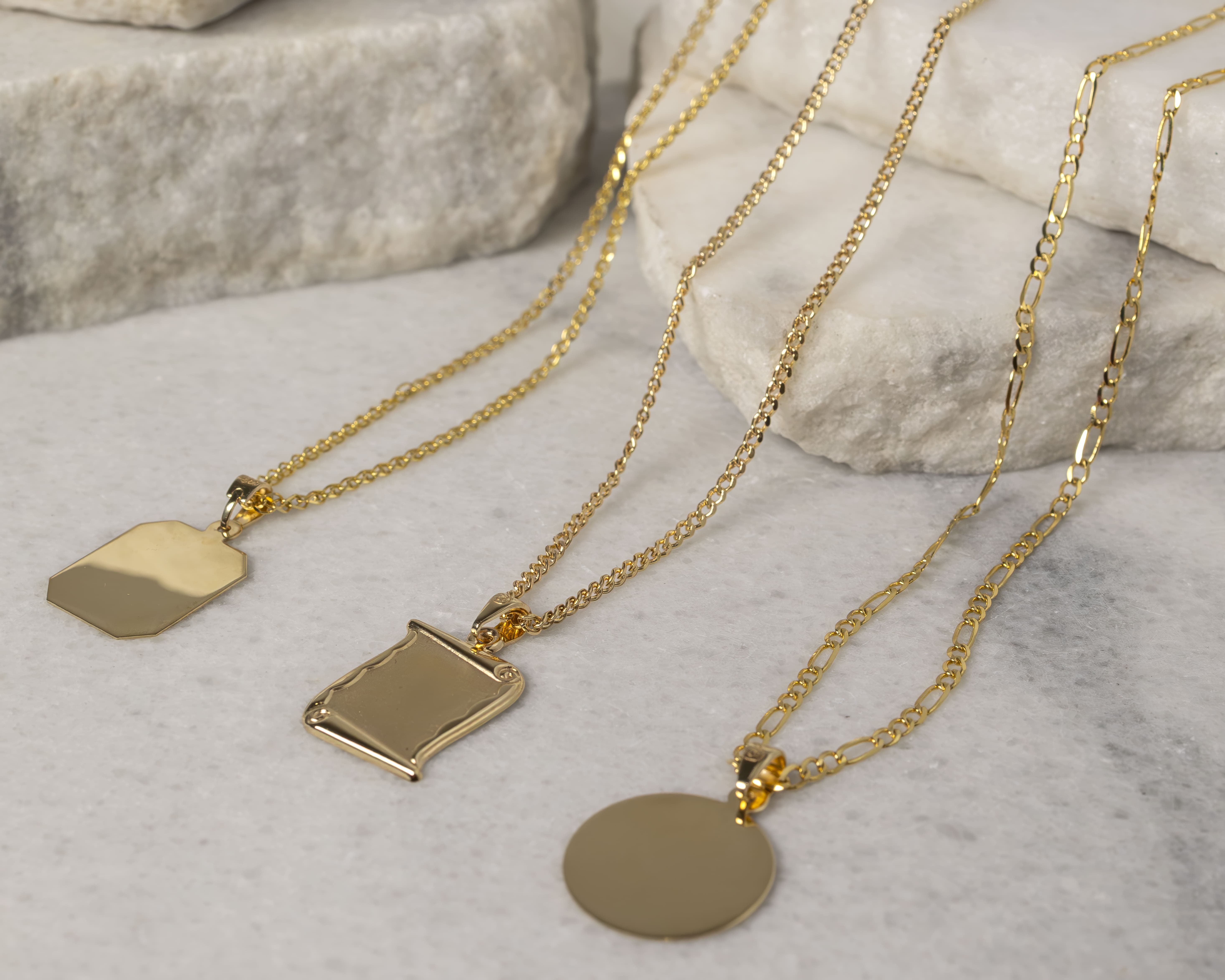 Three gold pendant necklaces with blank charms in shield, scroll, and round shapes displayed on white stone slabs.