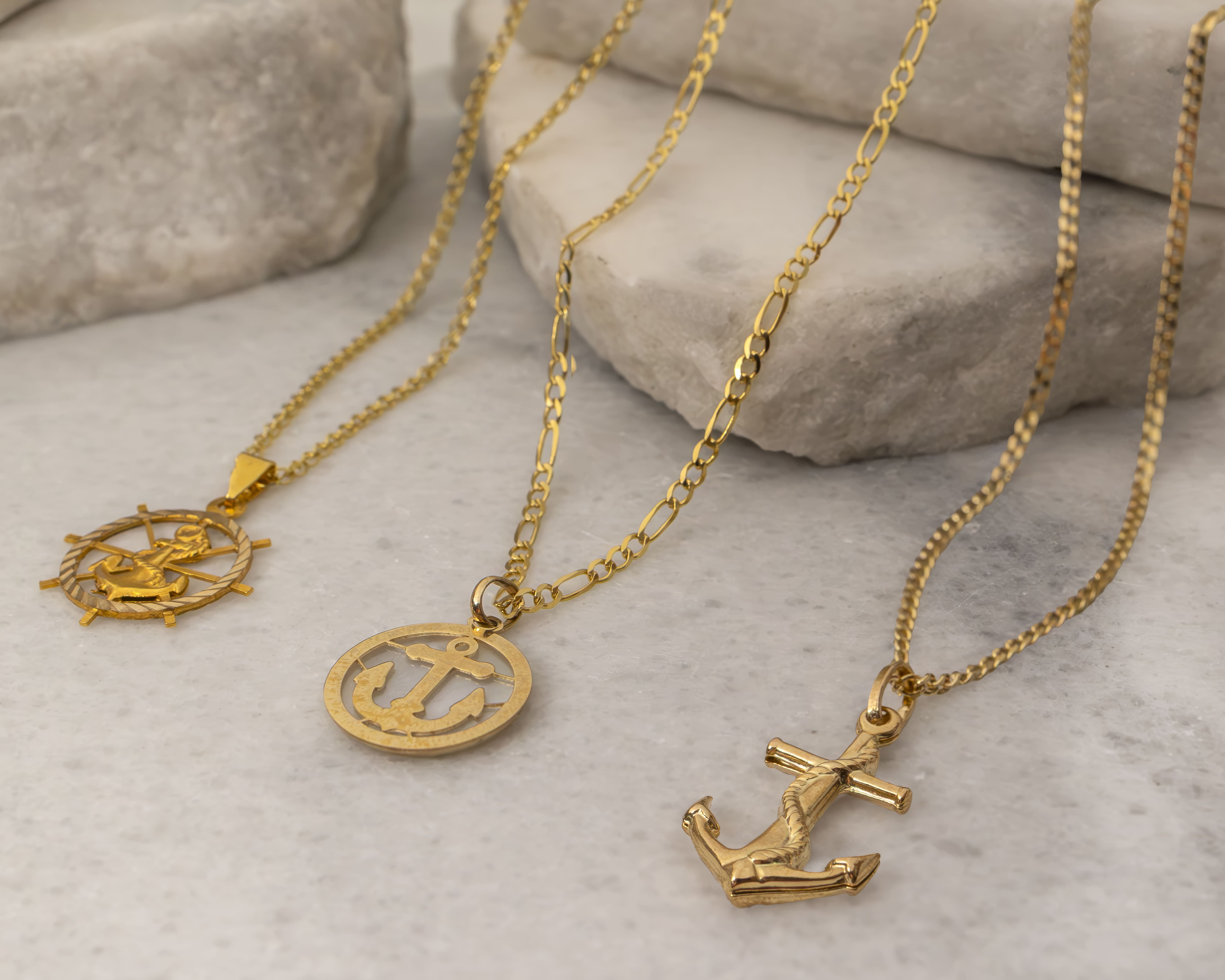 Three gold necklaces with maritime-themed pendants including an anchor and a ship wheel on a light stone surface.