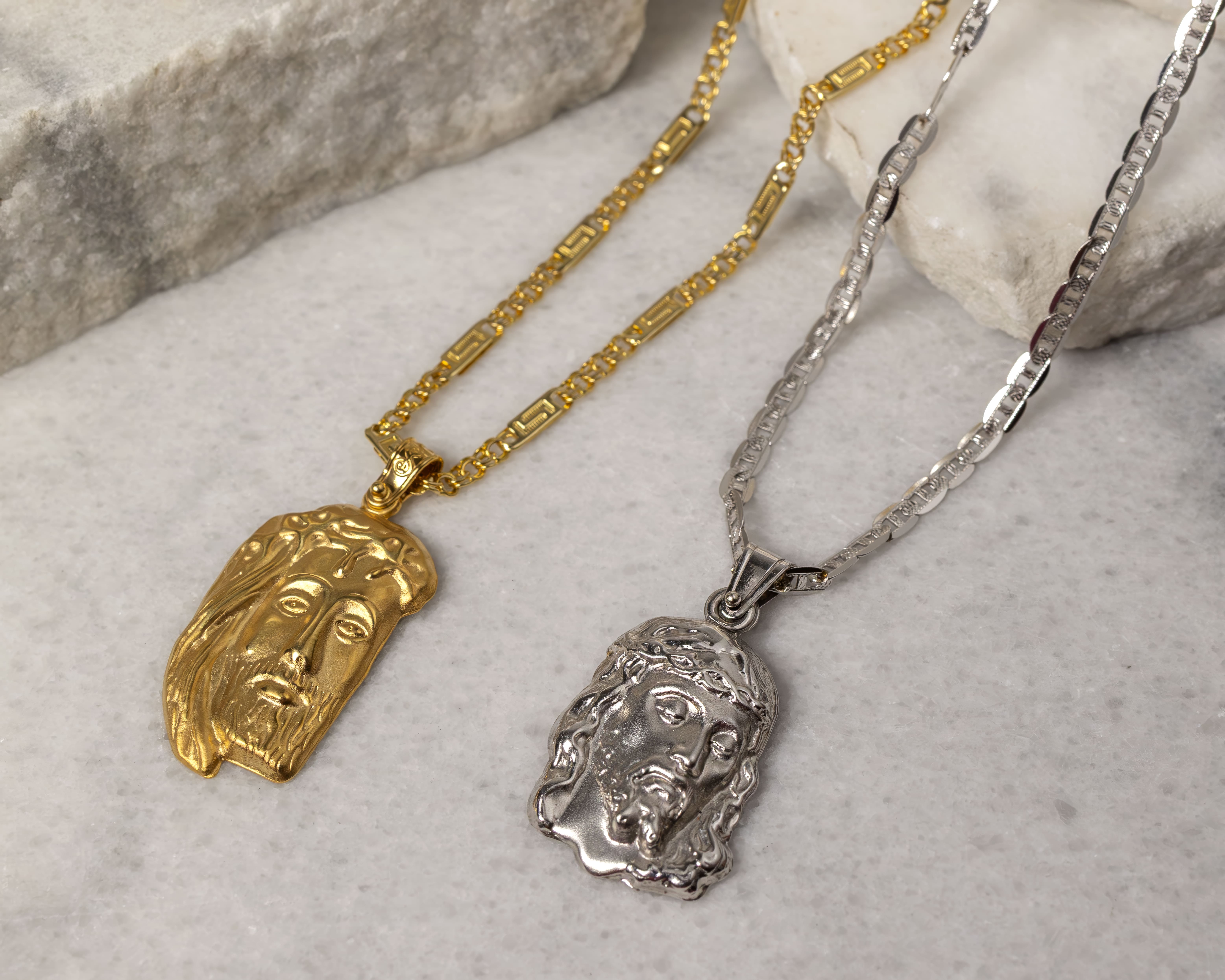 Two necklaces with pendants depicting Jesus Christ's face, one gold and one silver, displayed on a marble surface.
