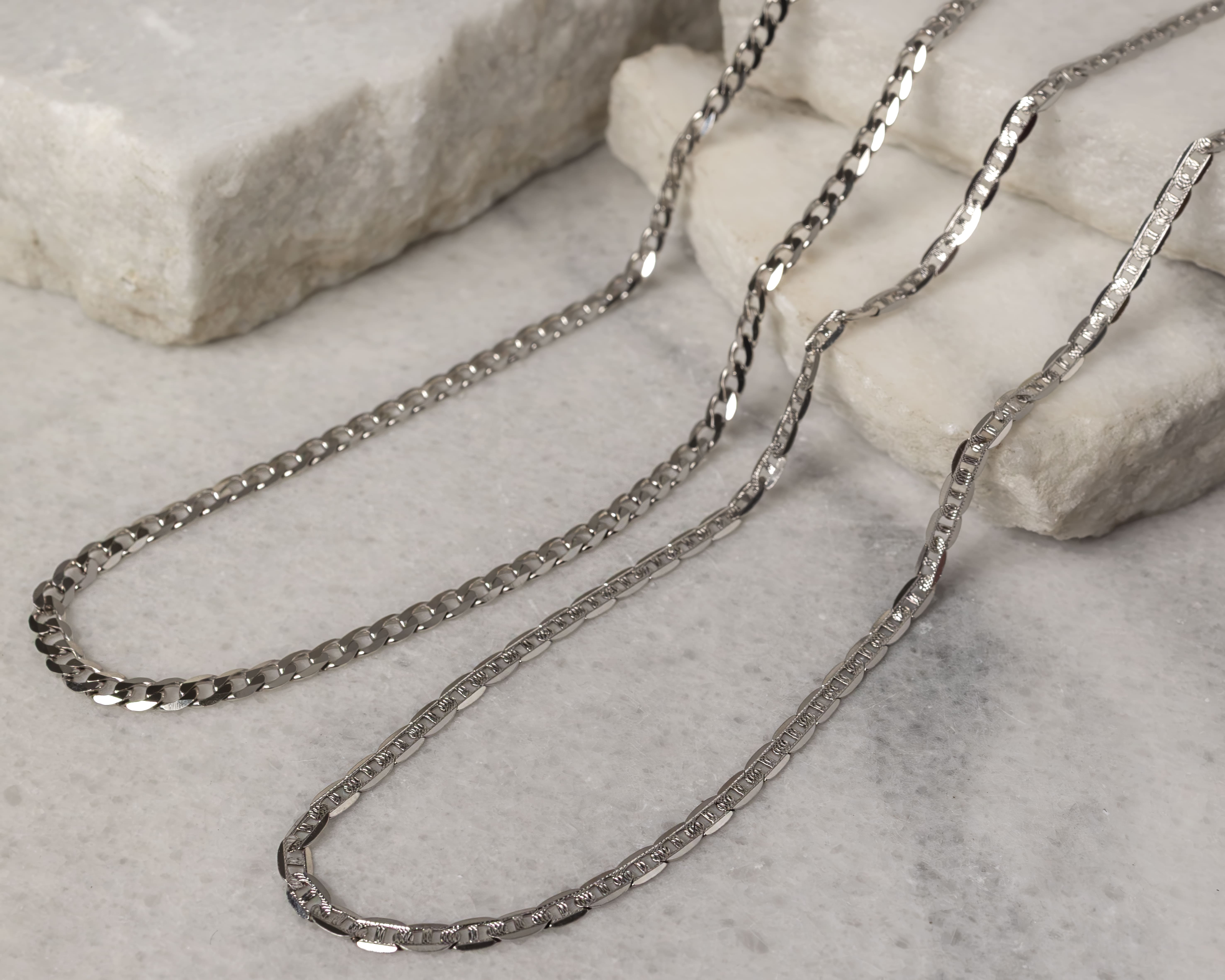 Two silver chains with different link patterns displayed on a light marble surface with white stone blocks.