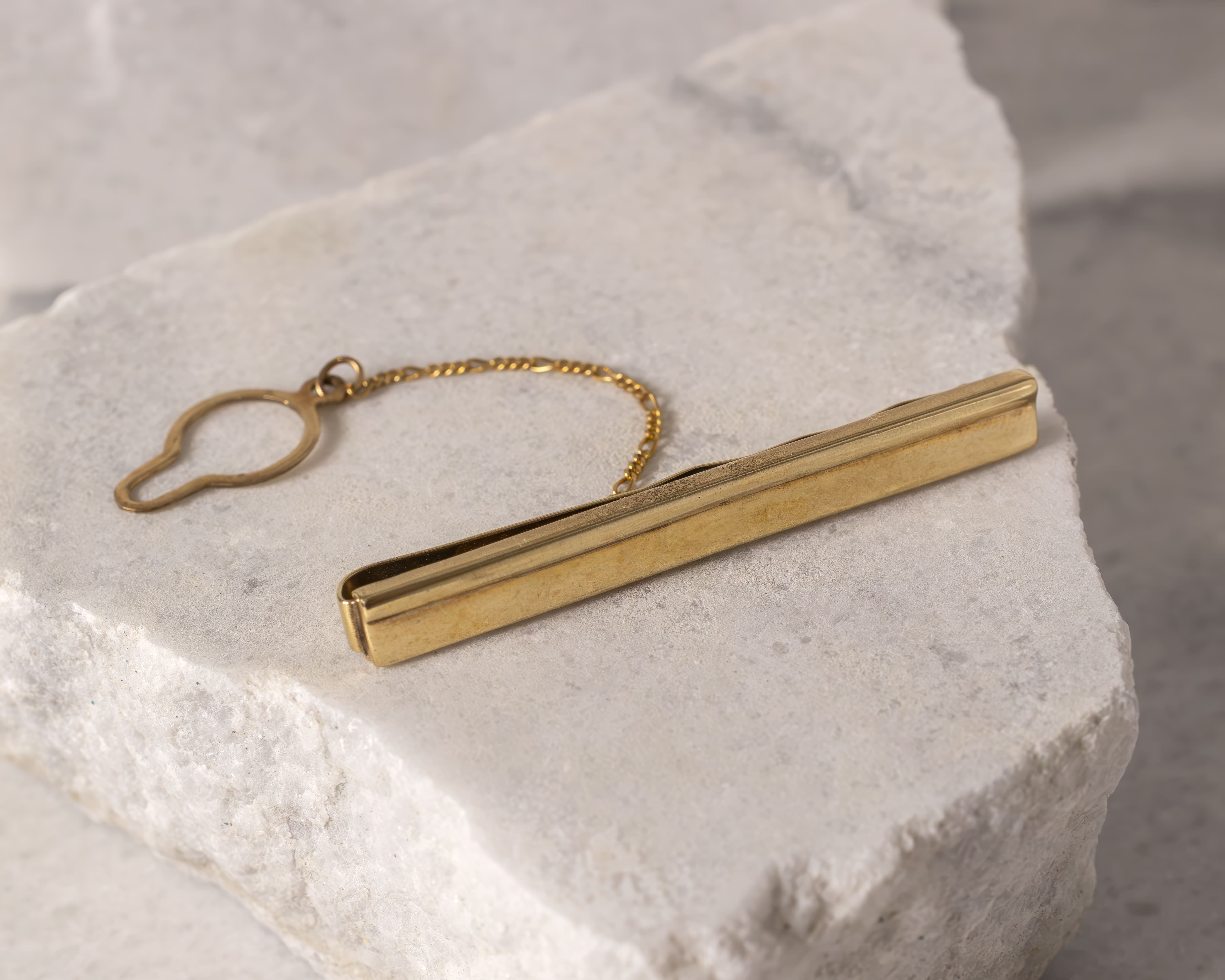 Gold-tone tie clip with attached chain and loop resting on rough white stone surface.