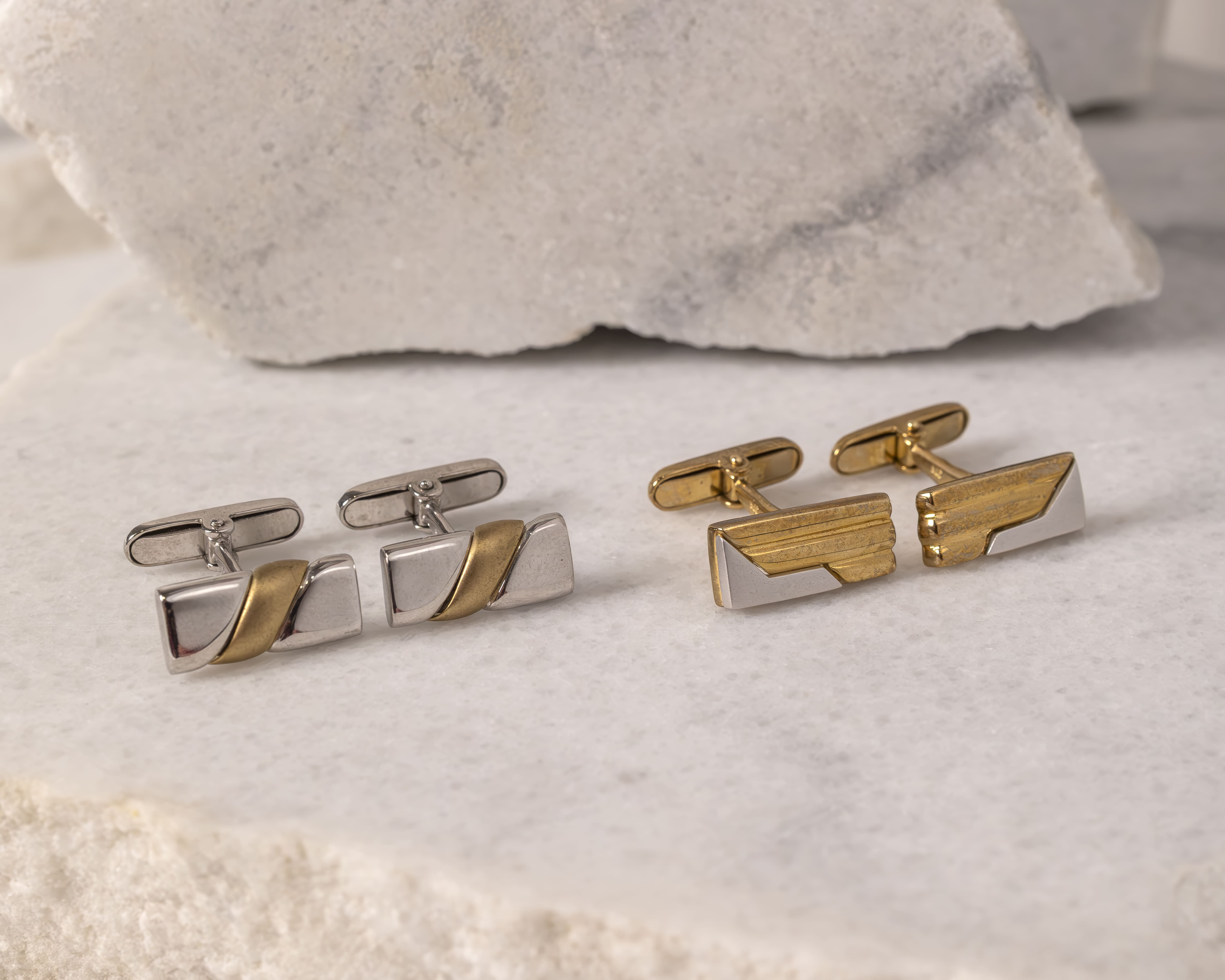 Two pairs of rectangular cufflinks on a marble surface, one pair silver with a gold diagonal stripe and the other gold with silver diagonal accents.