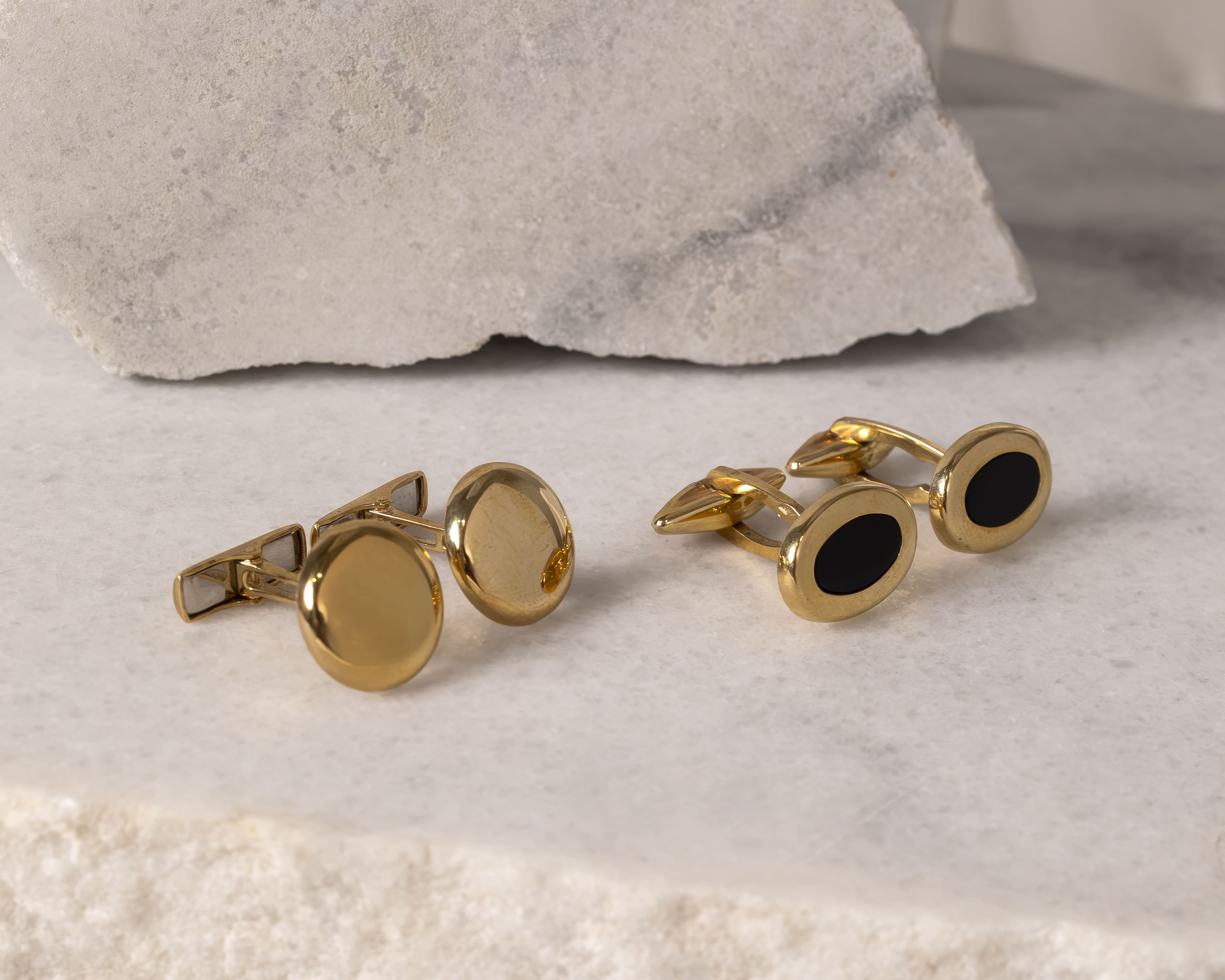 Two pairs of gold cufflinks, one pair with plain gold faces and the other with black oval insets, displayed on a stone surface.