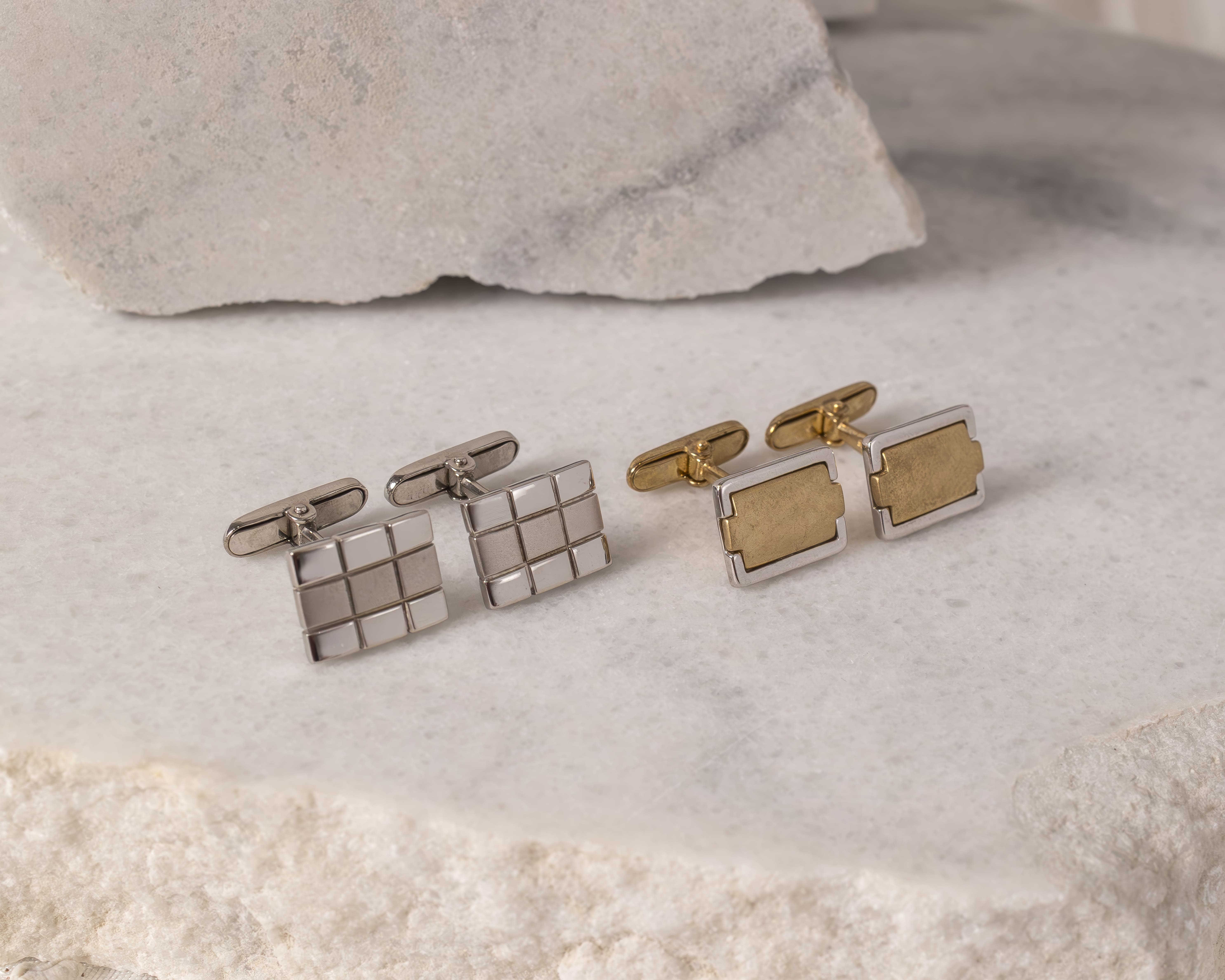 Two pairs of cufflinks on a white stone surface, one pair silver with a grid pattern and the other pair gold with a rectangular design.