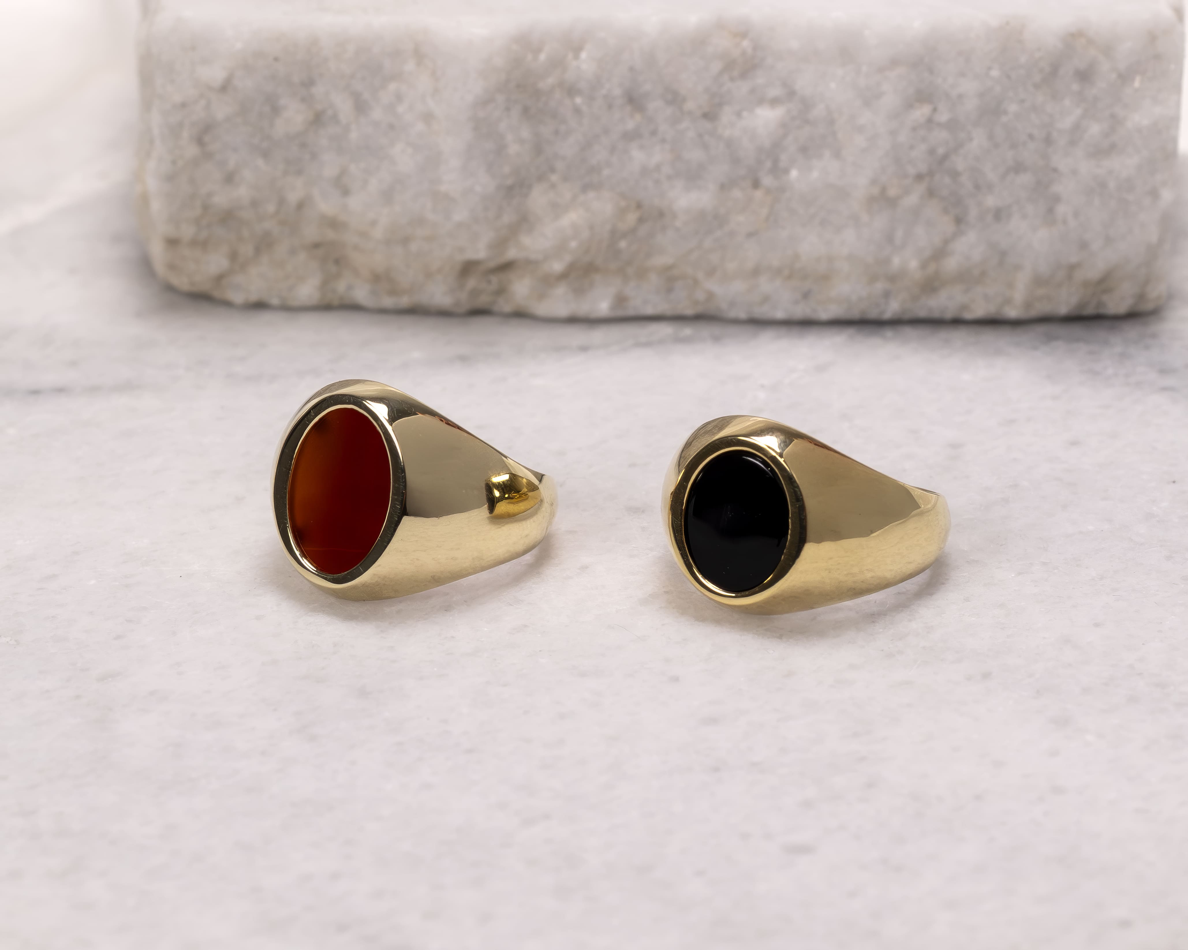 Two gold signet rings on a white marble surface; one with a red oval stone and the other with a black oval stone.