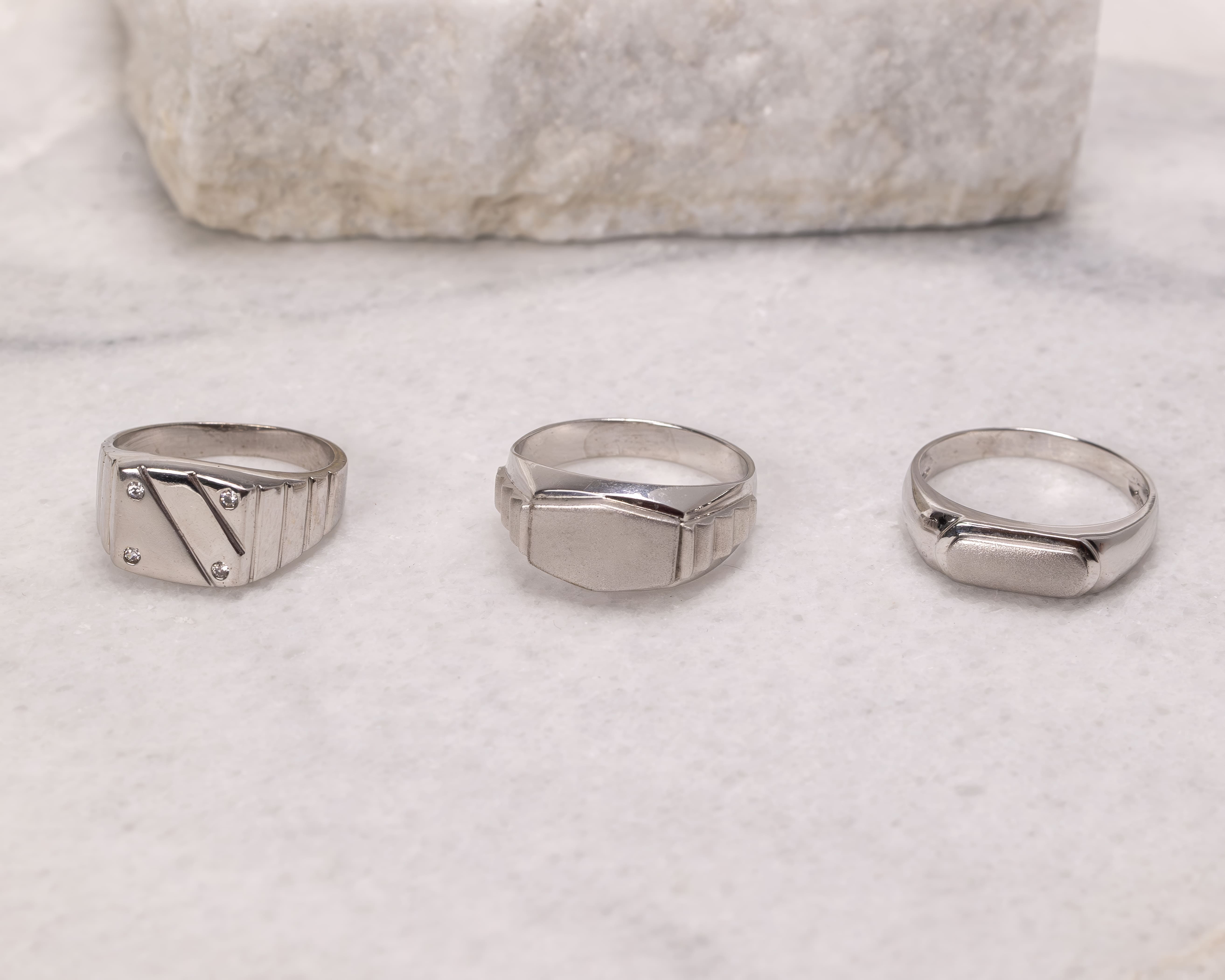 Three silver signet rings with different rectangular designs placed on a white marble surface.