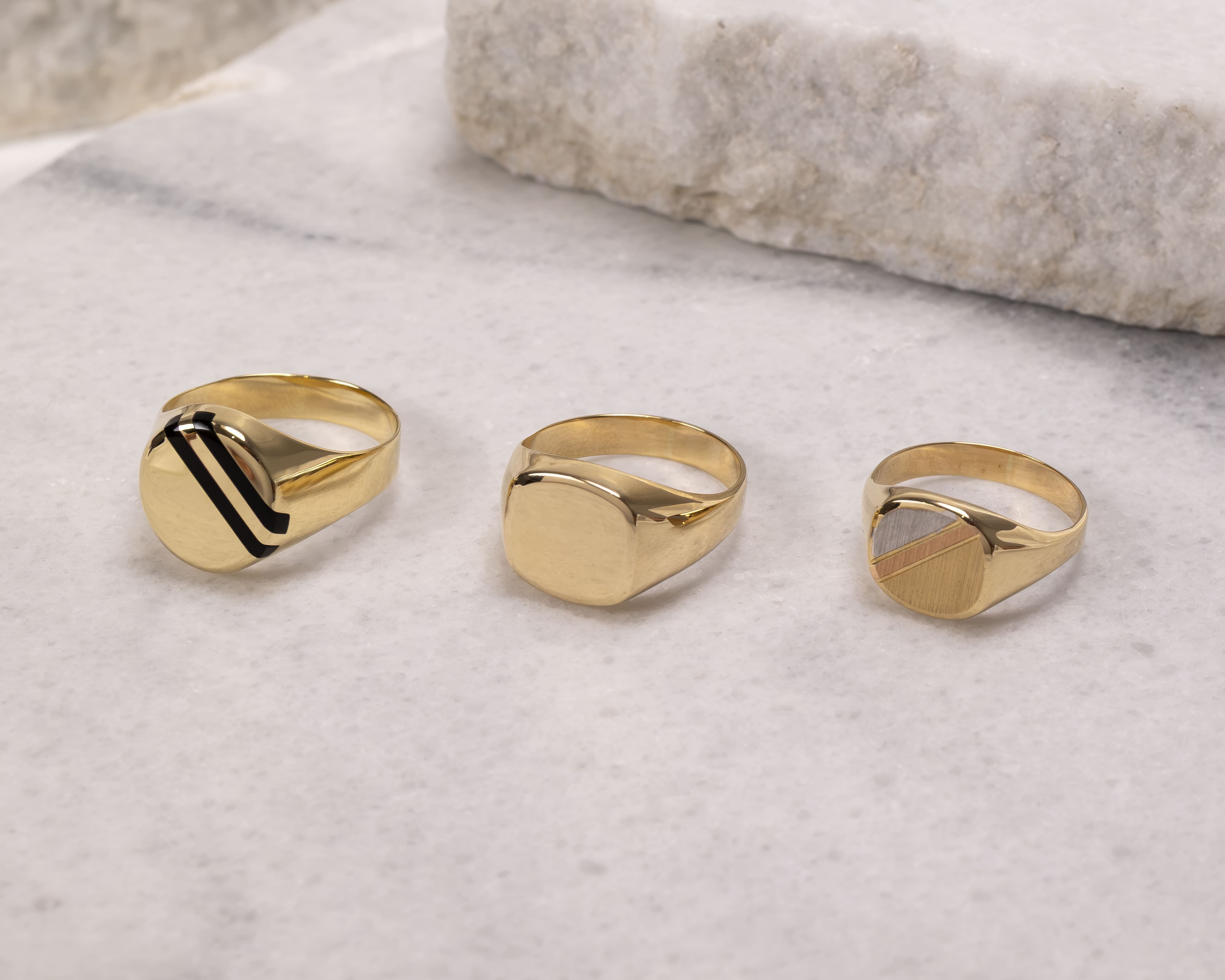 Three gold signet rings on a light marble surface, featuring varied plain and striped designs.