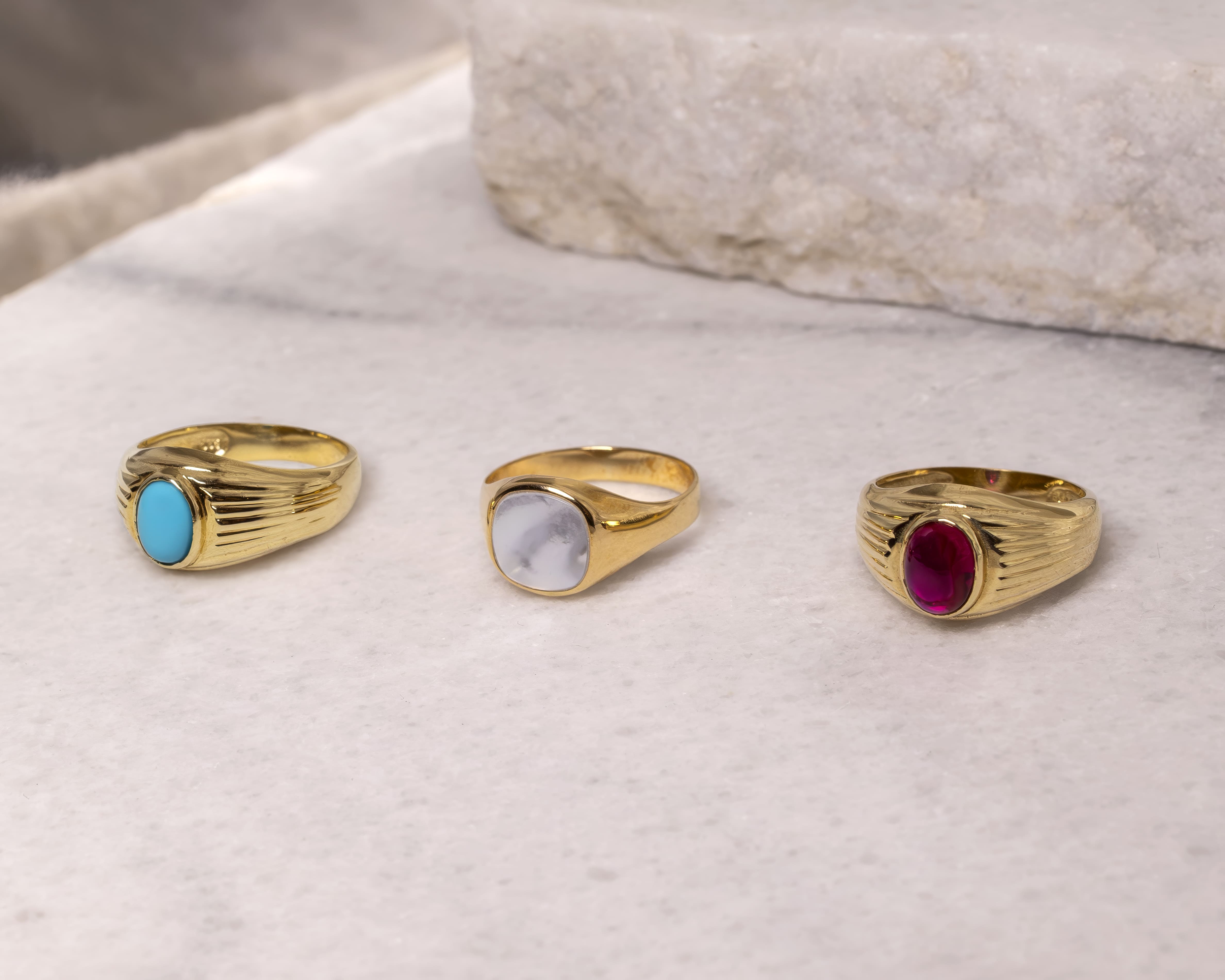 Three gold rings with oval stones: turquoise, white marbled, and deep red, displayed on a marble surface.