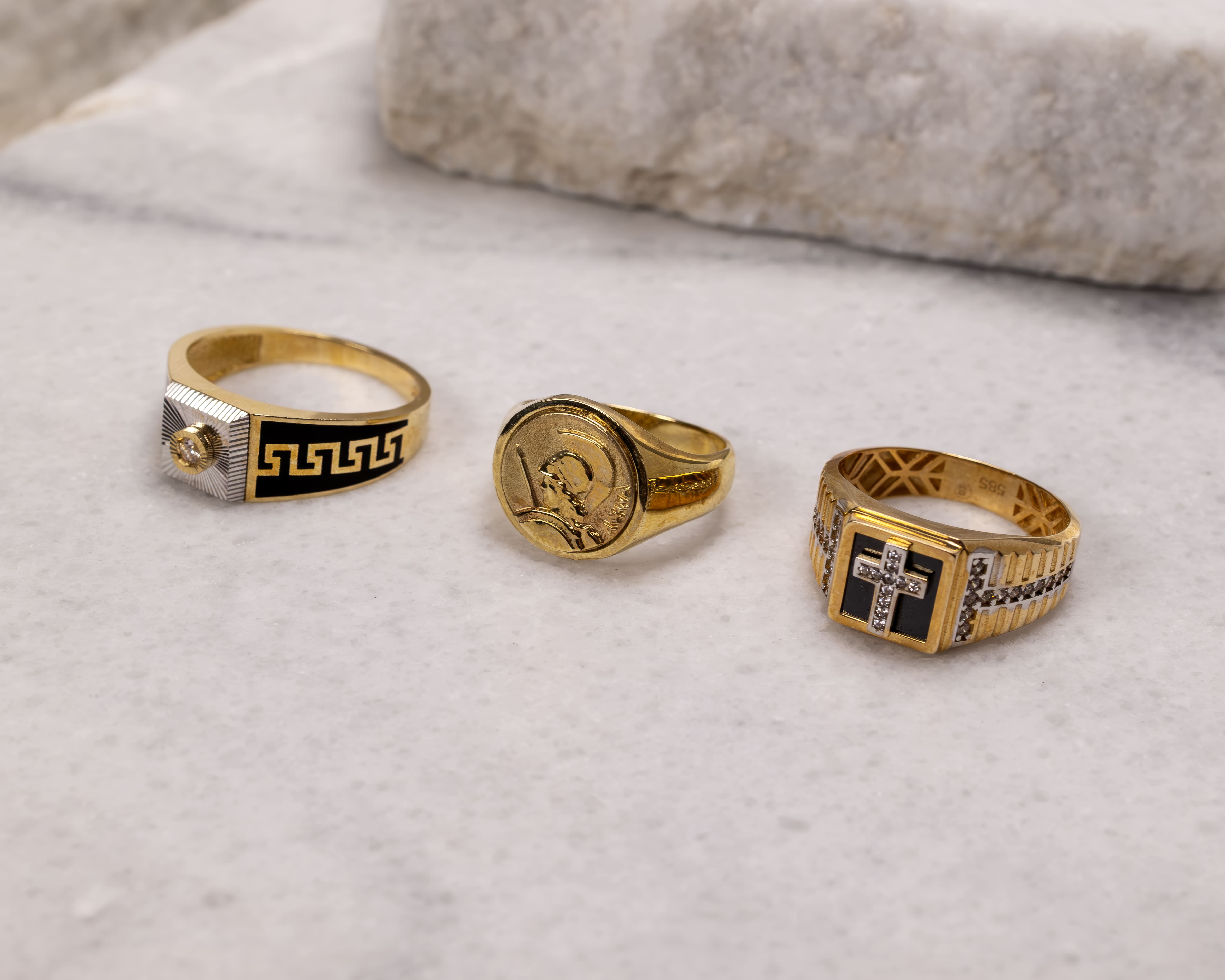 Three gold rings on a marble surface, featuring distinct designs including a Greek key pattern, a coin with a warrior profile, and a cross with embedded stones.