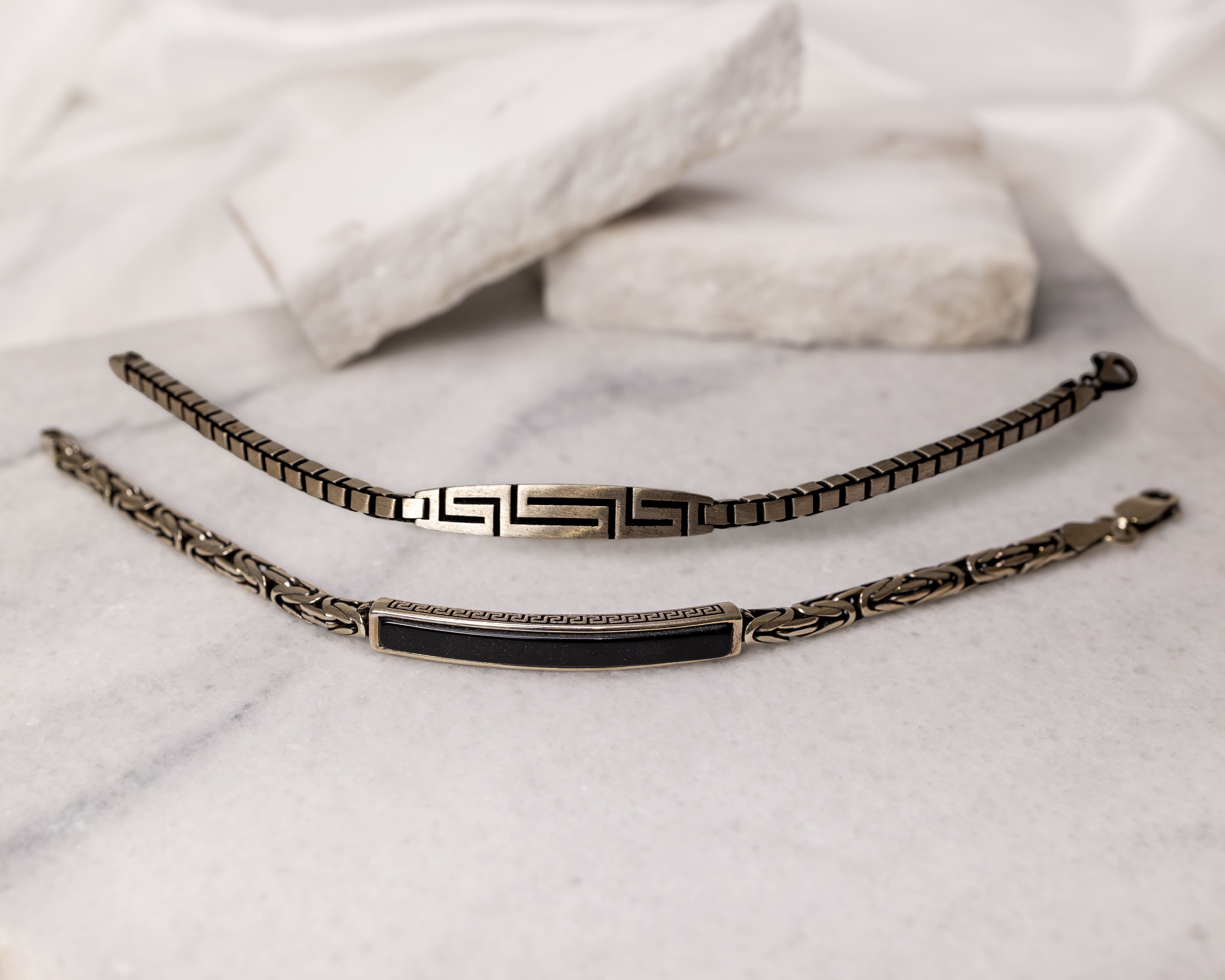 Two metallic bracelets with geometric and black inlay designs placed on a marble surface with blurred white stones in the background.