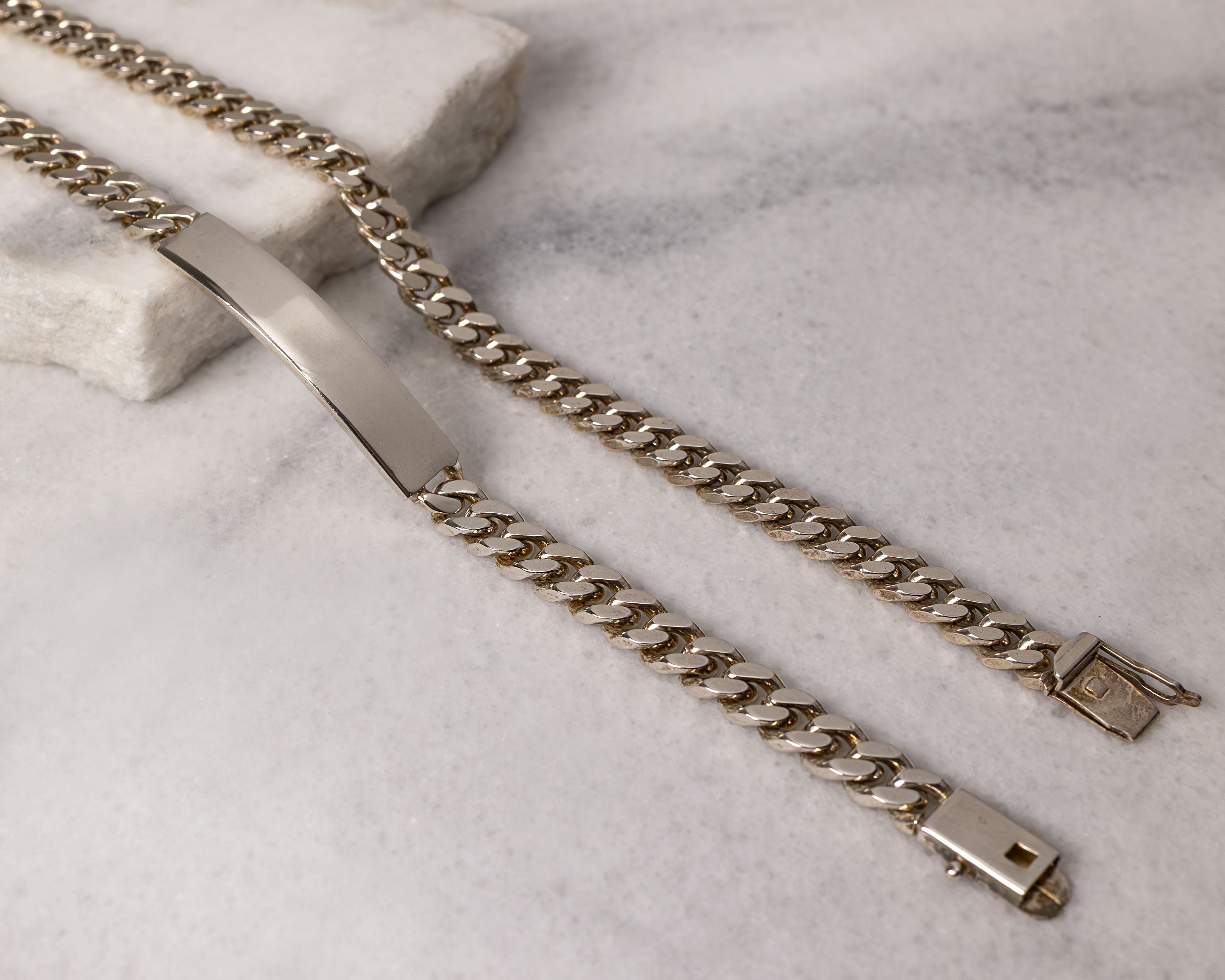 Silver curb chain bracelet with a blank rectangular ID plate lying open on a light marble surface.