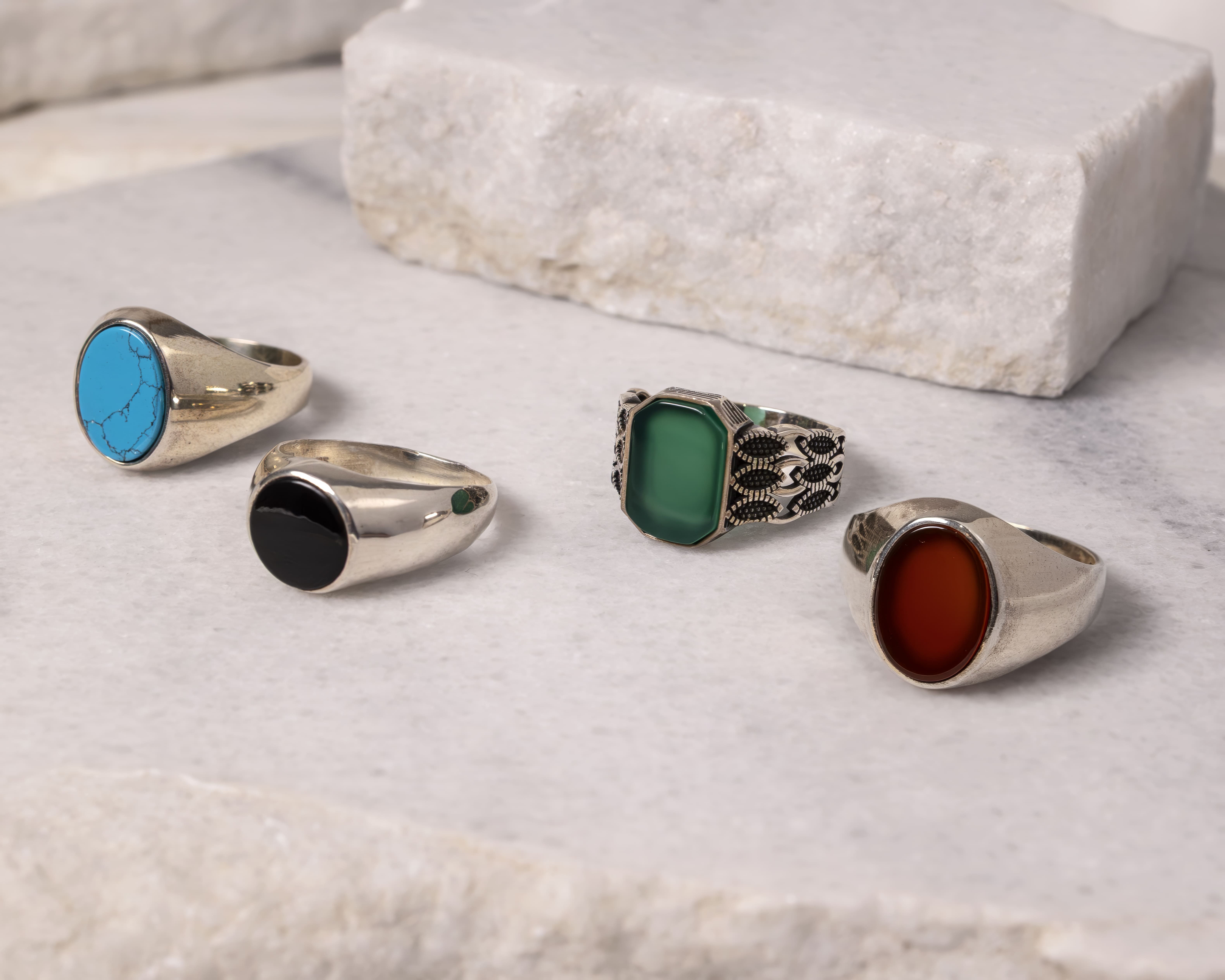 Four silver rings with different colored stones: turquoise, black, green, and reddish-brown, displayed on a light marble surface.