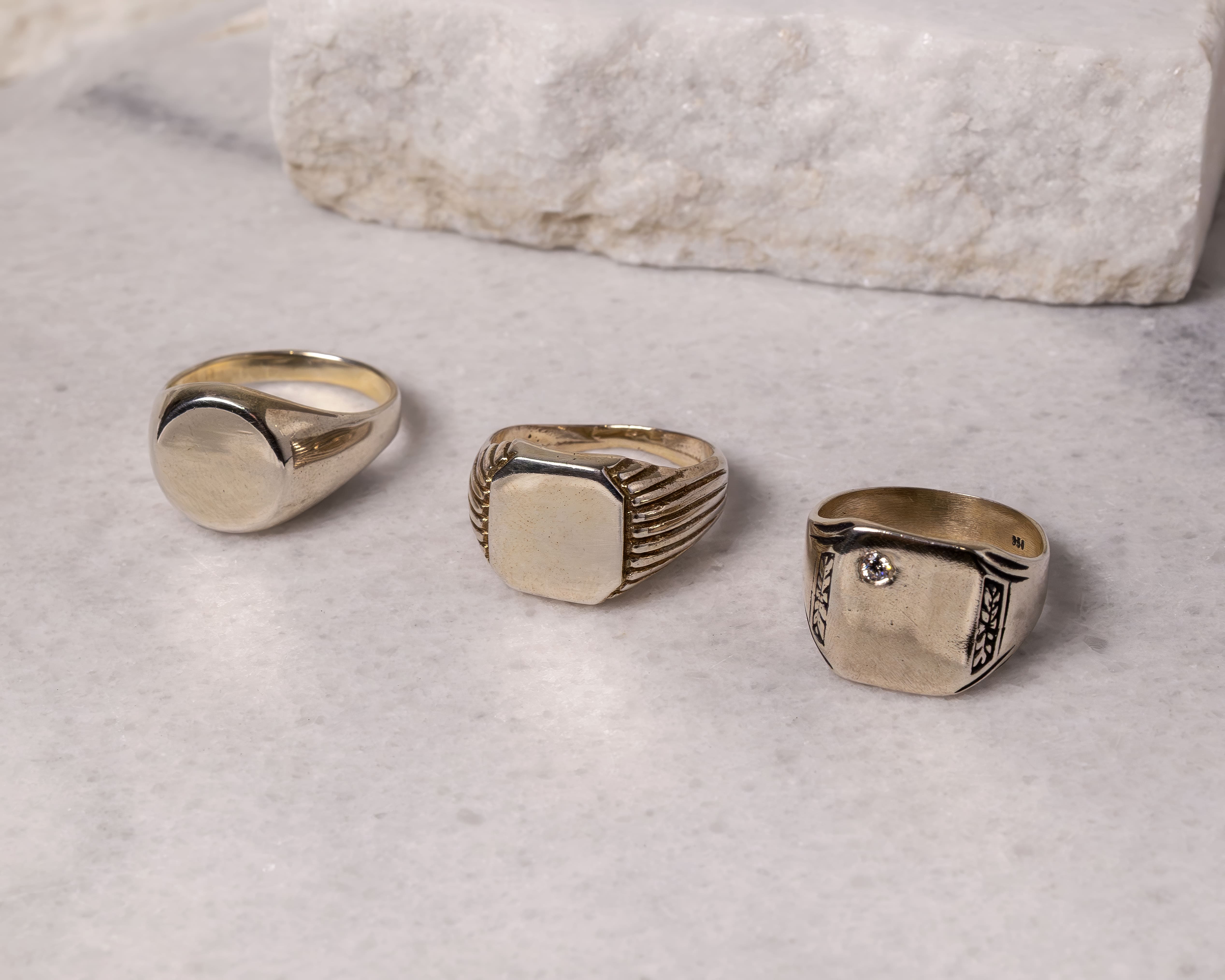 Three gold signet rings with different designs displayed on a white marble surface.