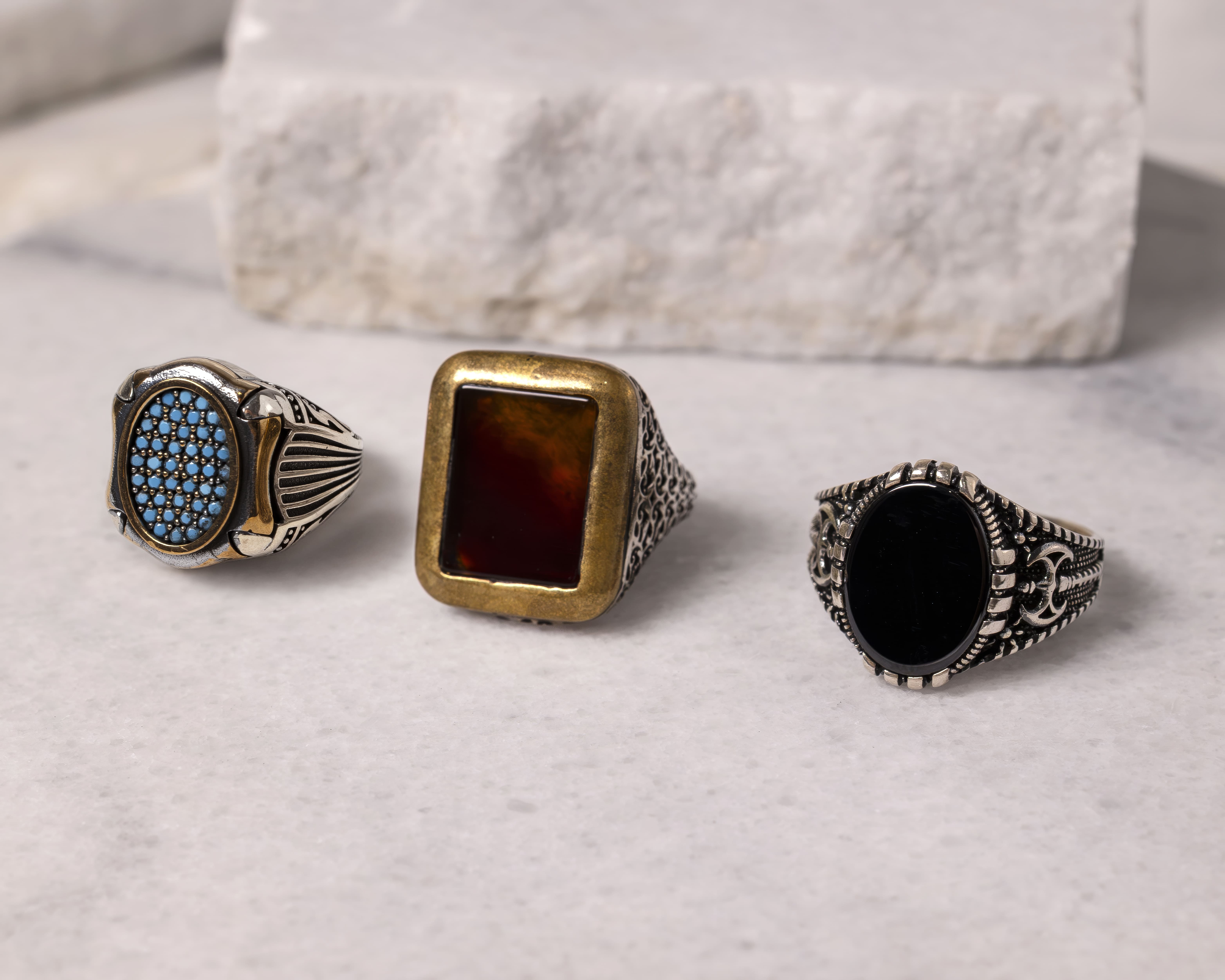 Three ornate men's rings with different stones: one with small blue beads, one with a rectangular amber-colored stone, and one with a solid black oval stone.