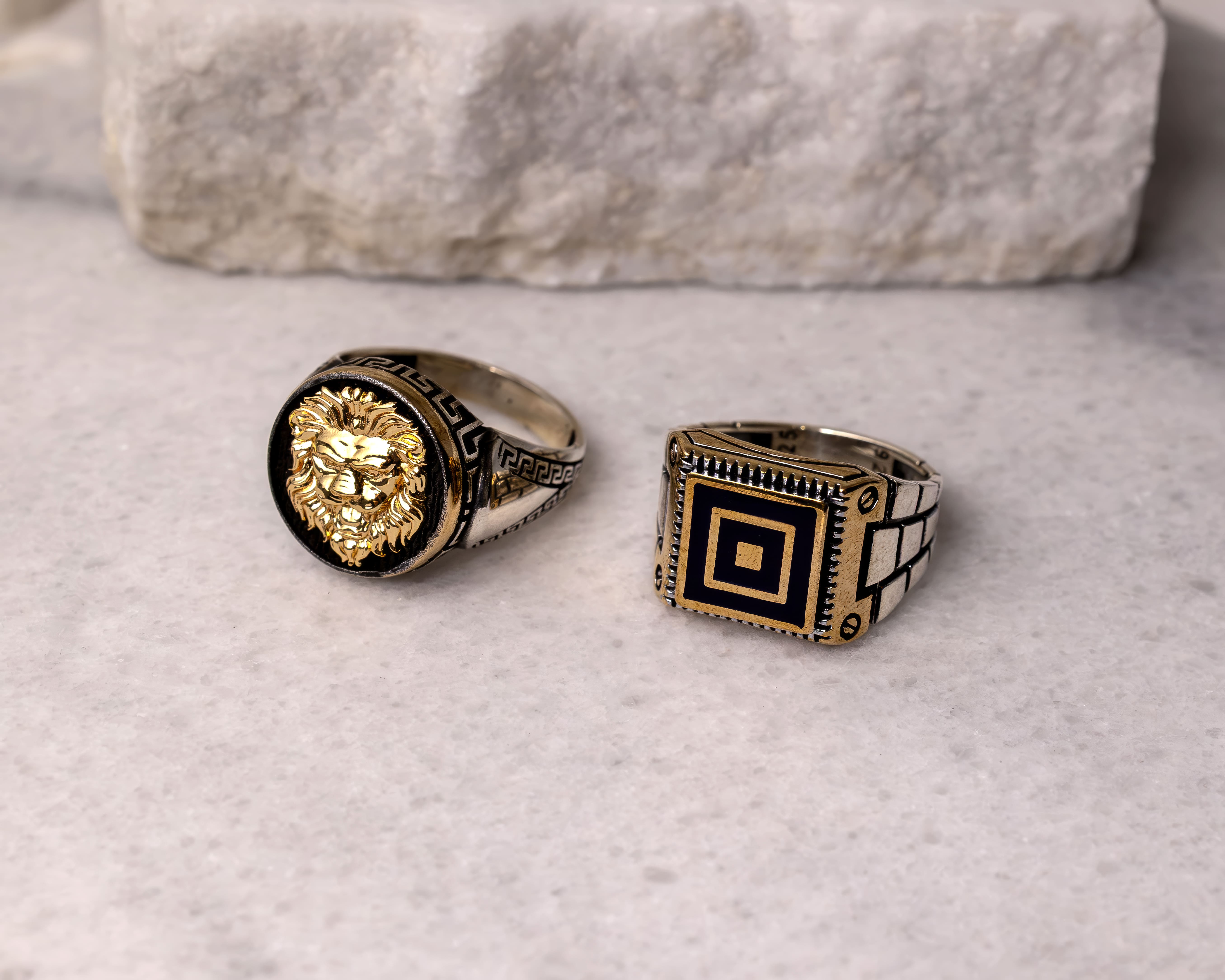Two silver and gold men's rings on a marble surface; one with a lion head design and the other with a square geometric pattern.