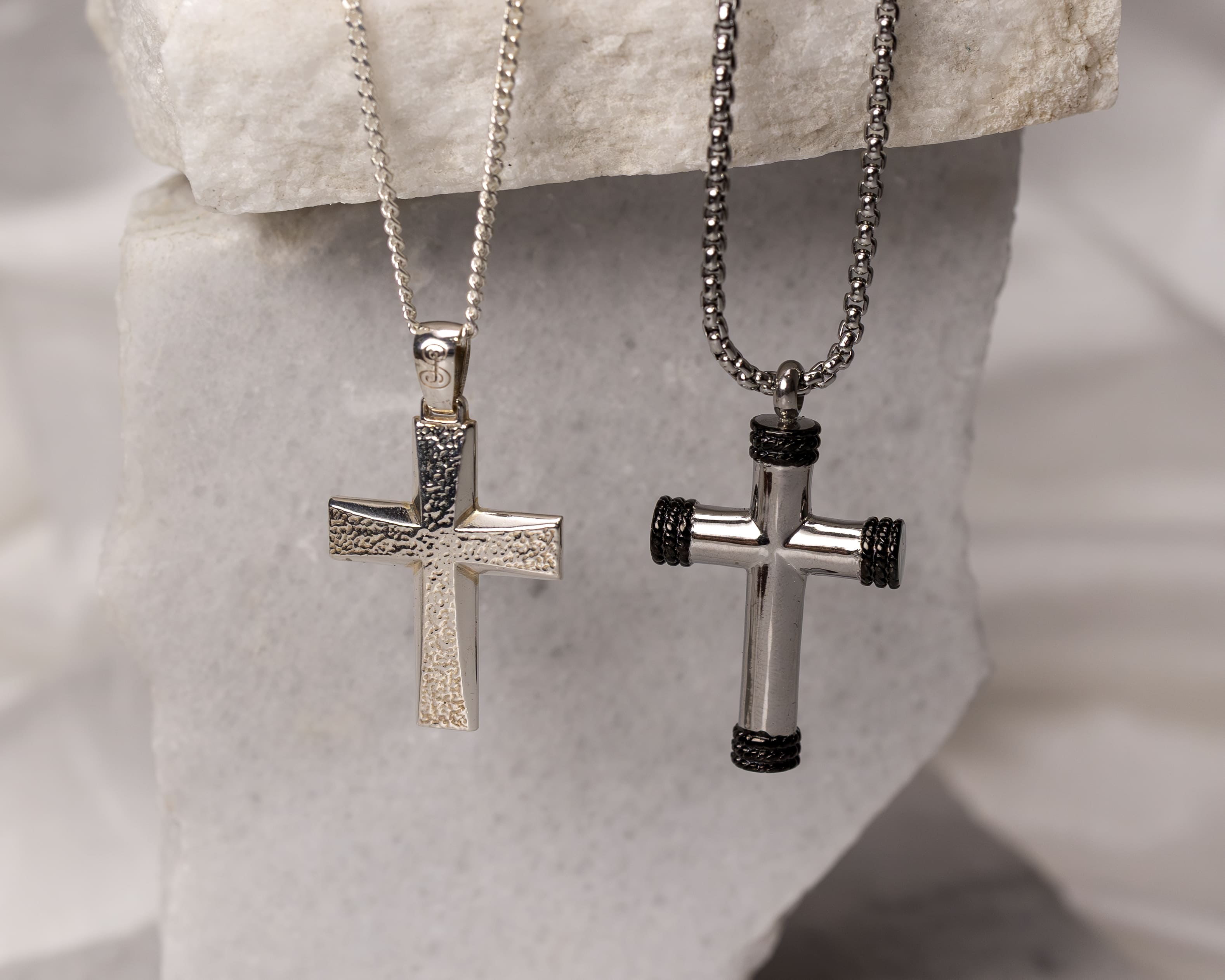 Two silver cross pendants hanging on chains against a textured stone background; one cross has a hammered texture while the other has black rope-like accents on its ends.