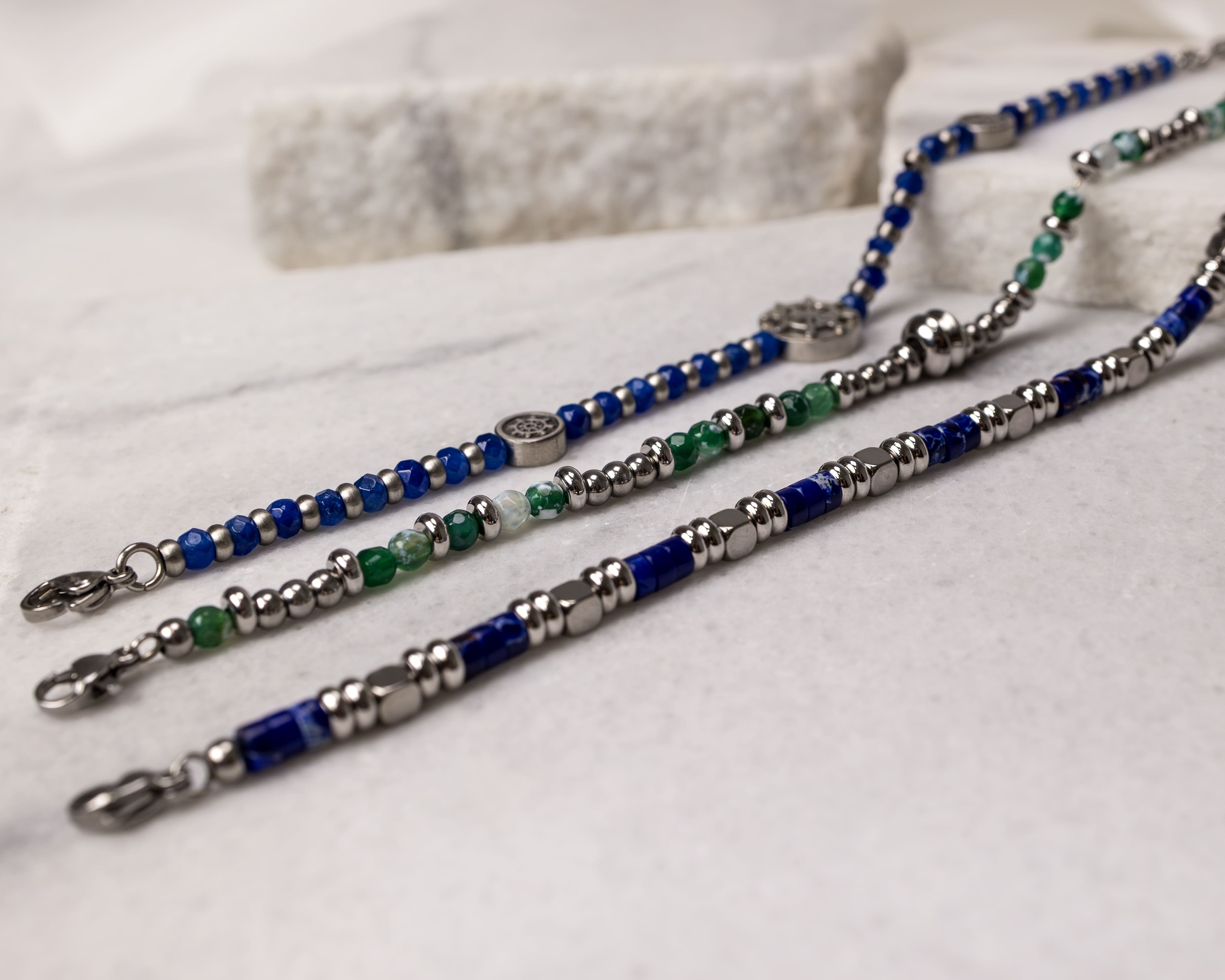 Three beaded bracelets with blue, green, and silver beads laid on a marble surface.