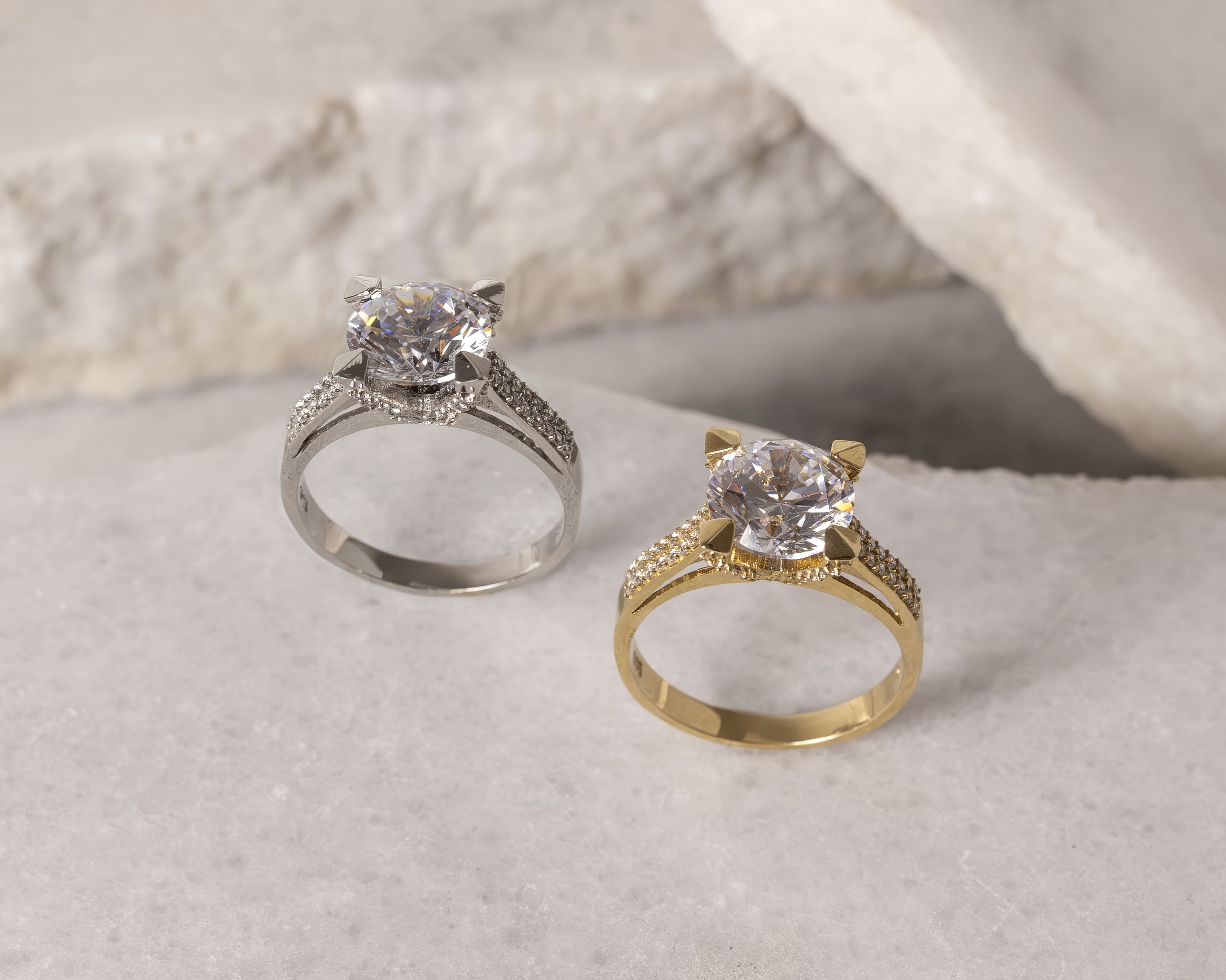 Two solitaire engagement rings with large round clear stones; one silver and one gold, placed on a light marble surface.