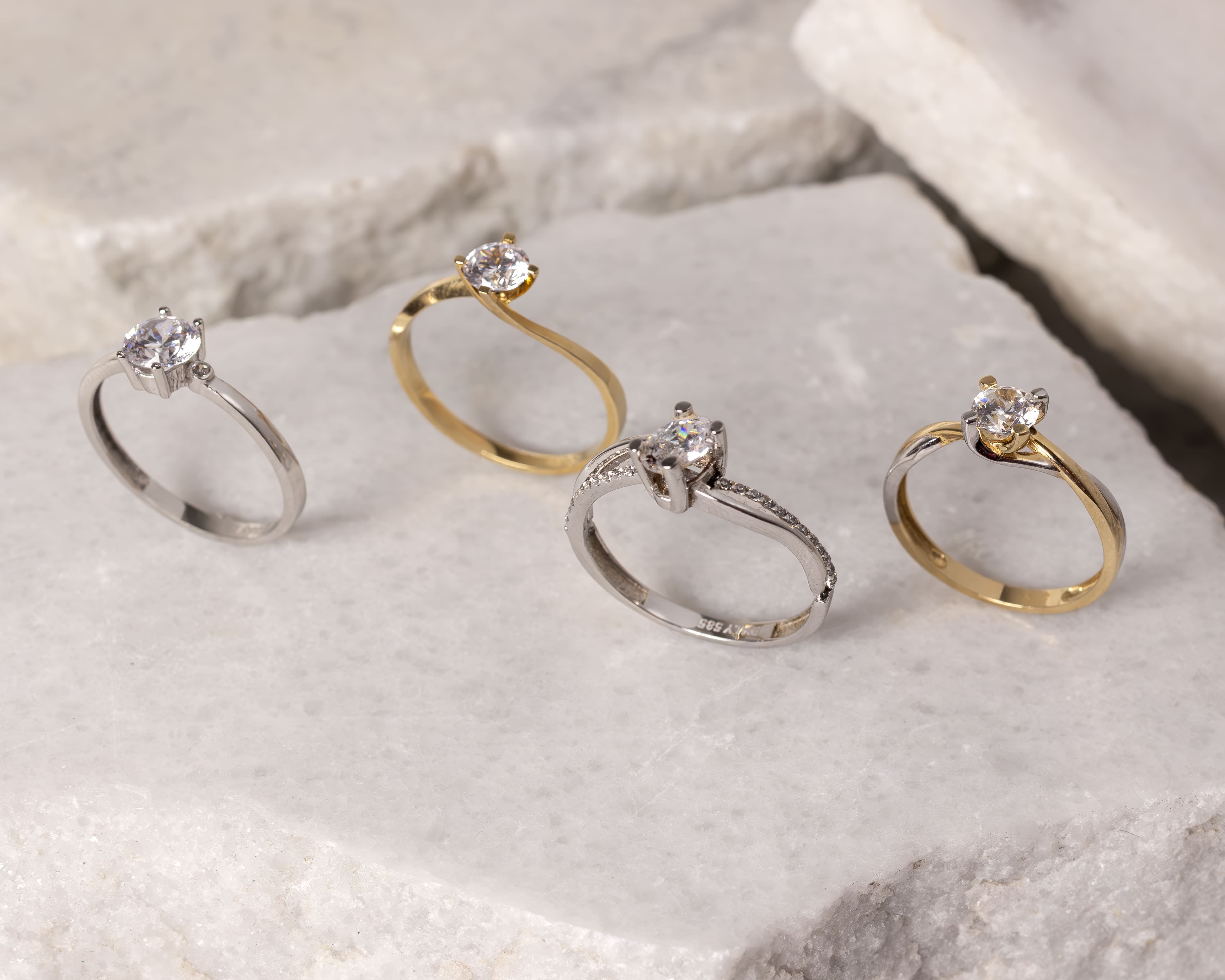 Four solitaire rings with clear round gemstones, two in gold and two in silver, displayed on a white stone surface.