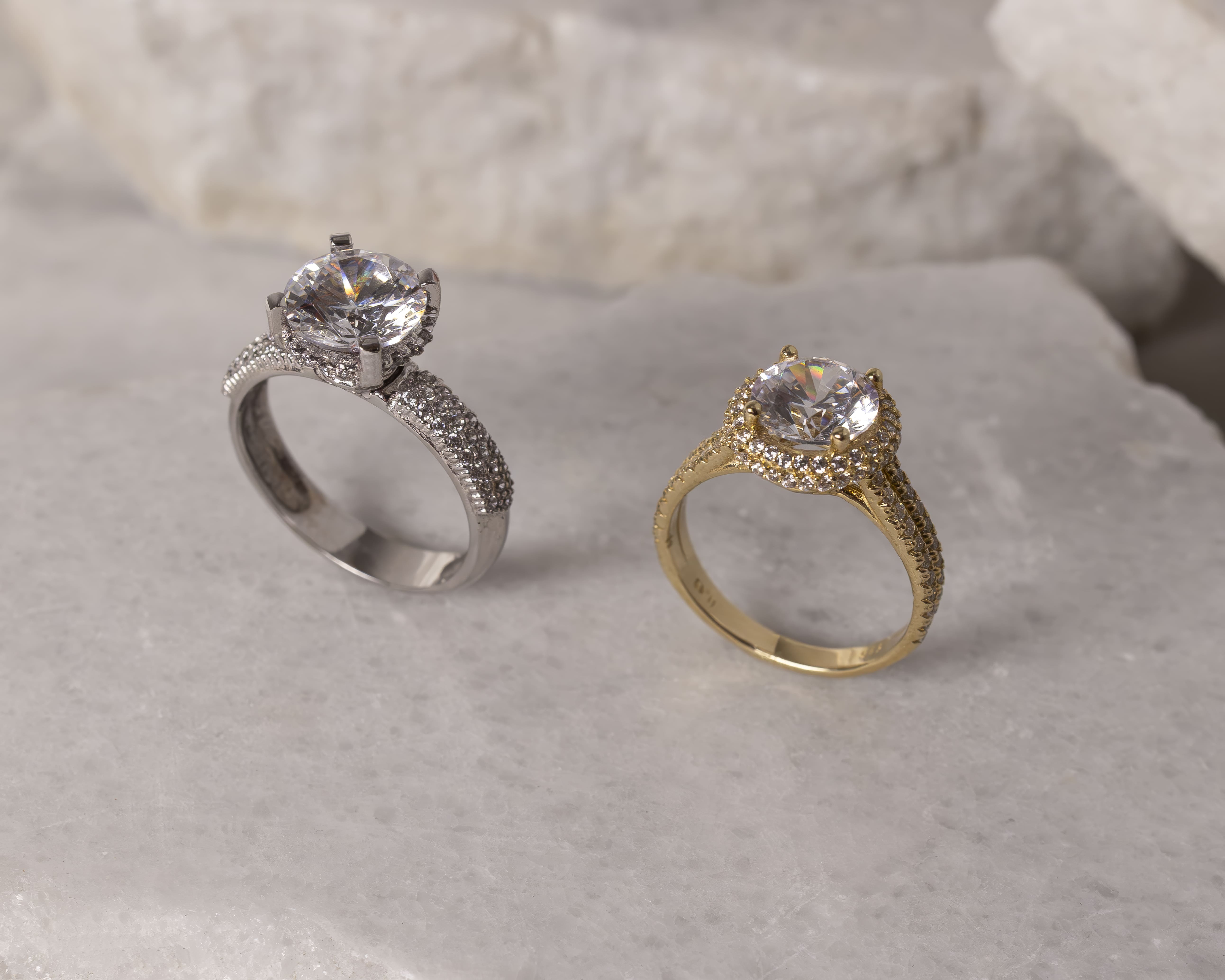 Close-up of two diamond engagement rings, one silver and one gold, displayed on a light marble surface.