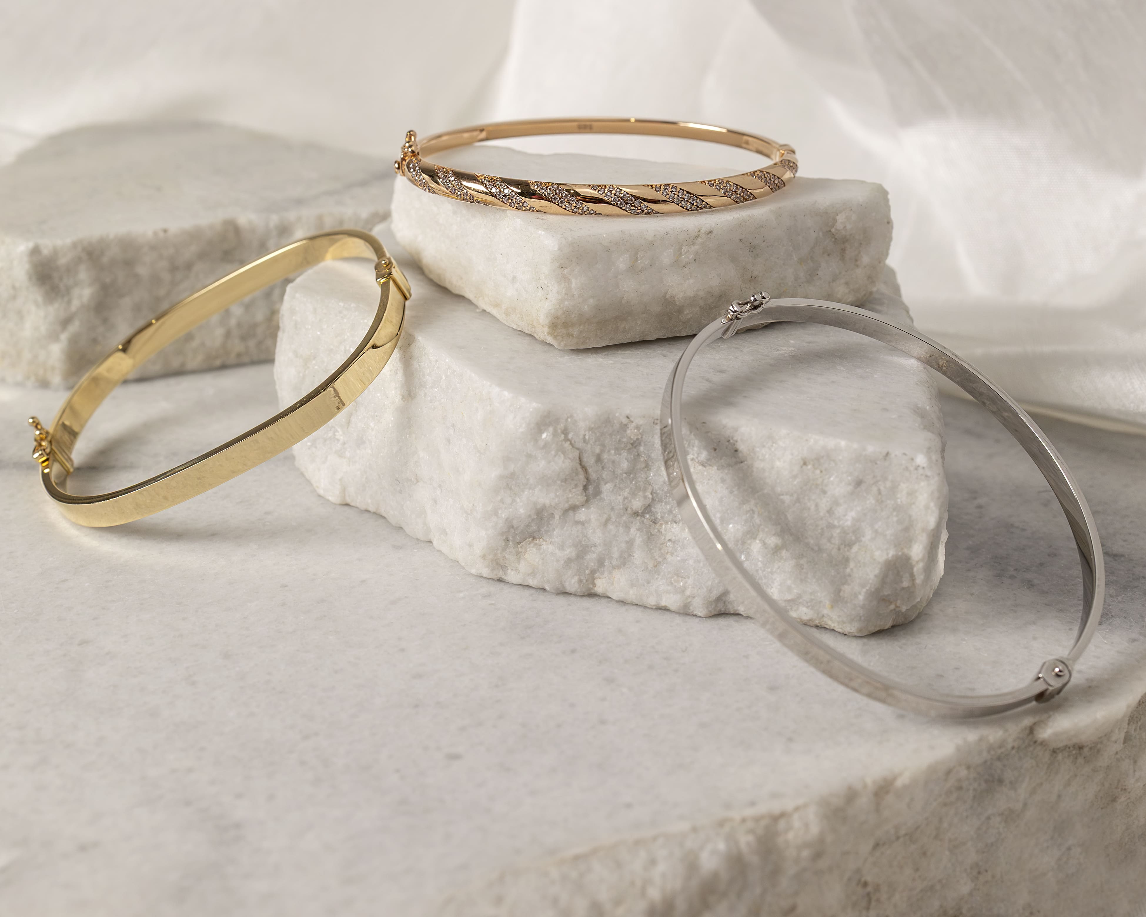 Three metal bangle bracelets in gold, rose gold with diamonds, and silver displayed on white marble blocks.