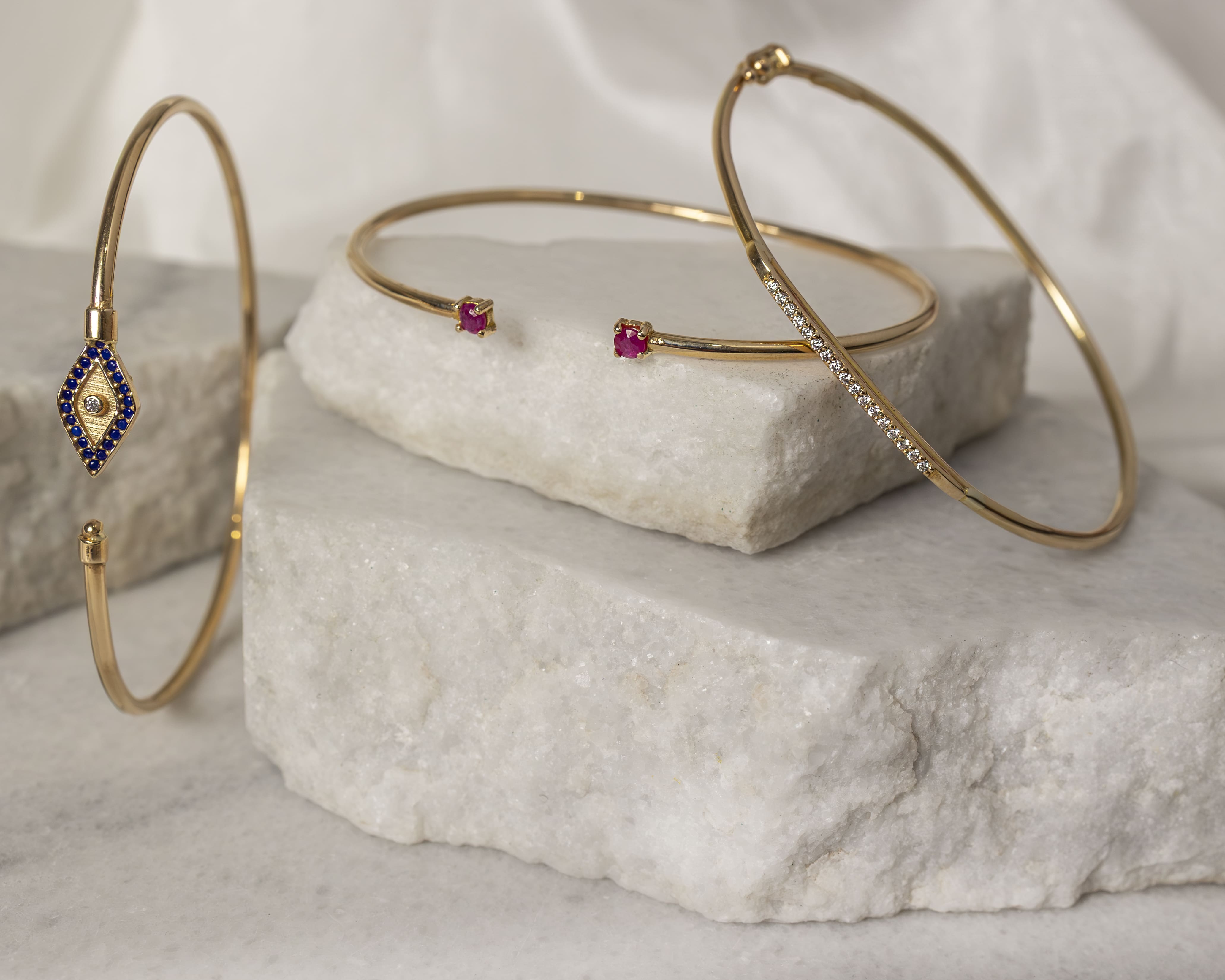Three delicate gold bracelets displayed on white marble slabs, featuring blue stones, red ruby ends, and small white diamonds.