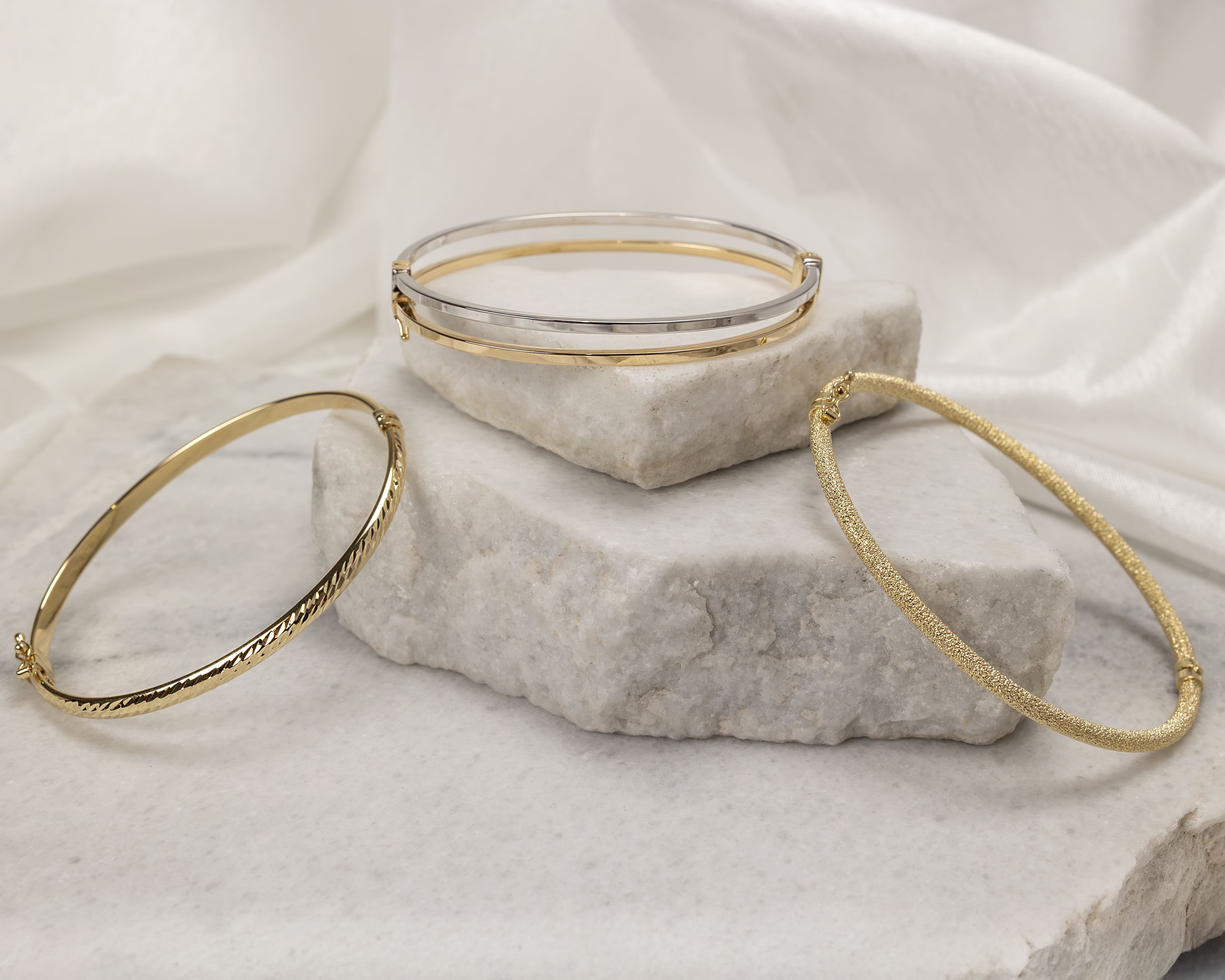 Three gold and silver bracelets displayed on stacked white marble slabs with a soft white fabric background.