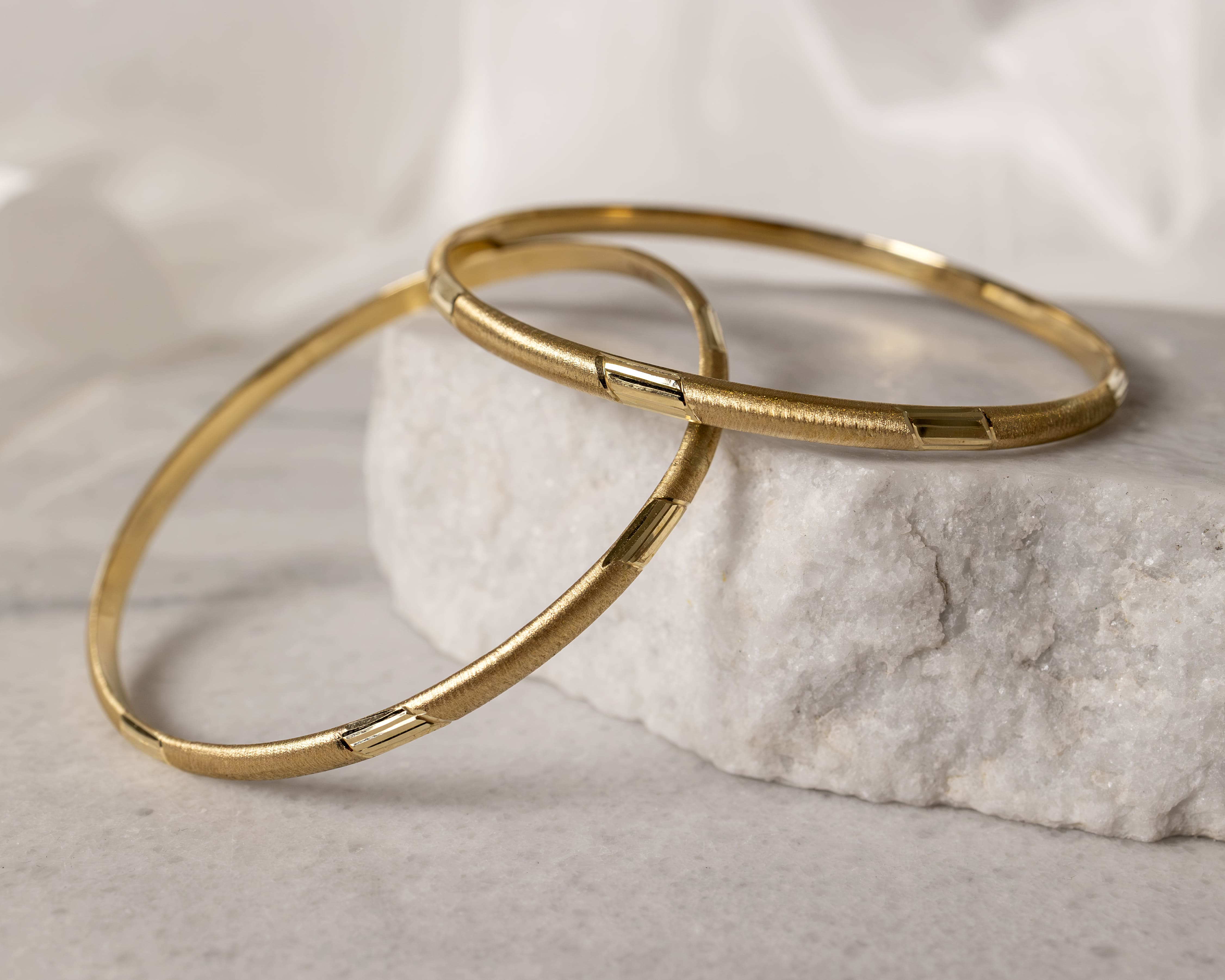 Two textured gold bangles resting on a white stone surface with a soft white background.