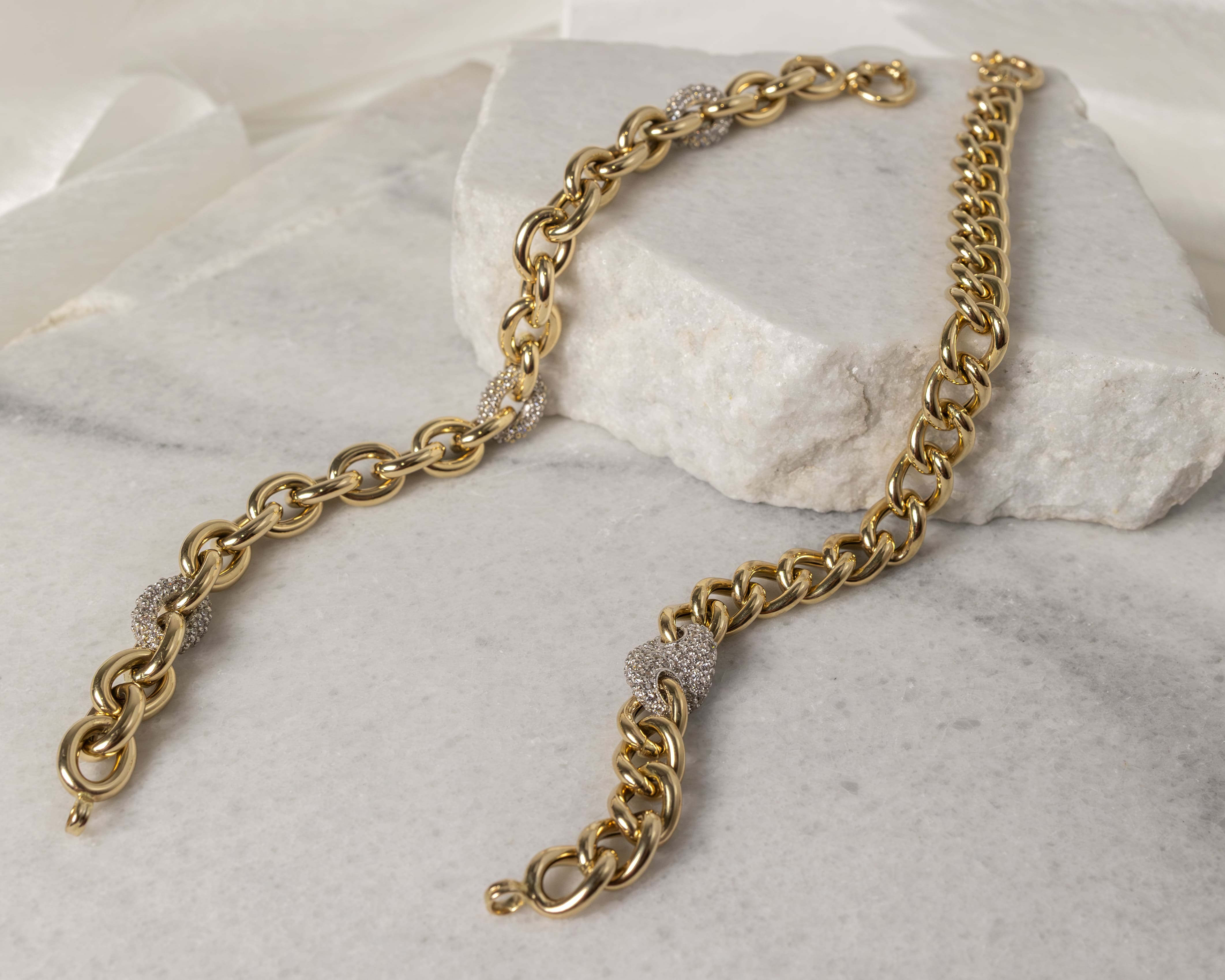Two gold chain bracelets with diamond-encrusted links displayed on white marble slabs.