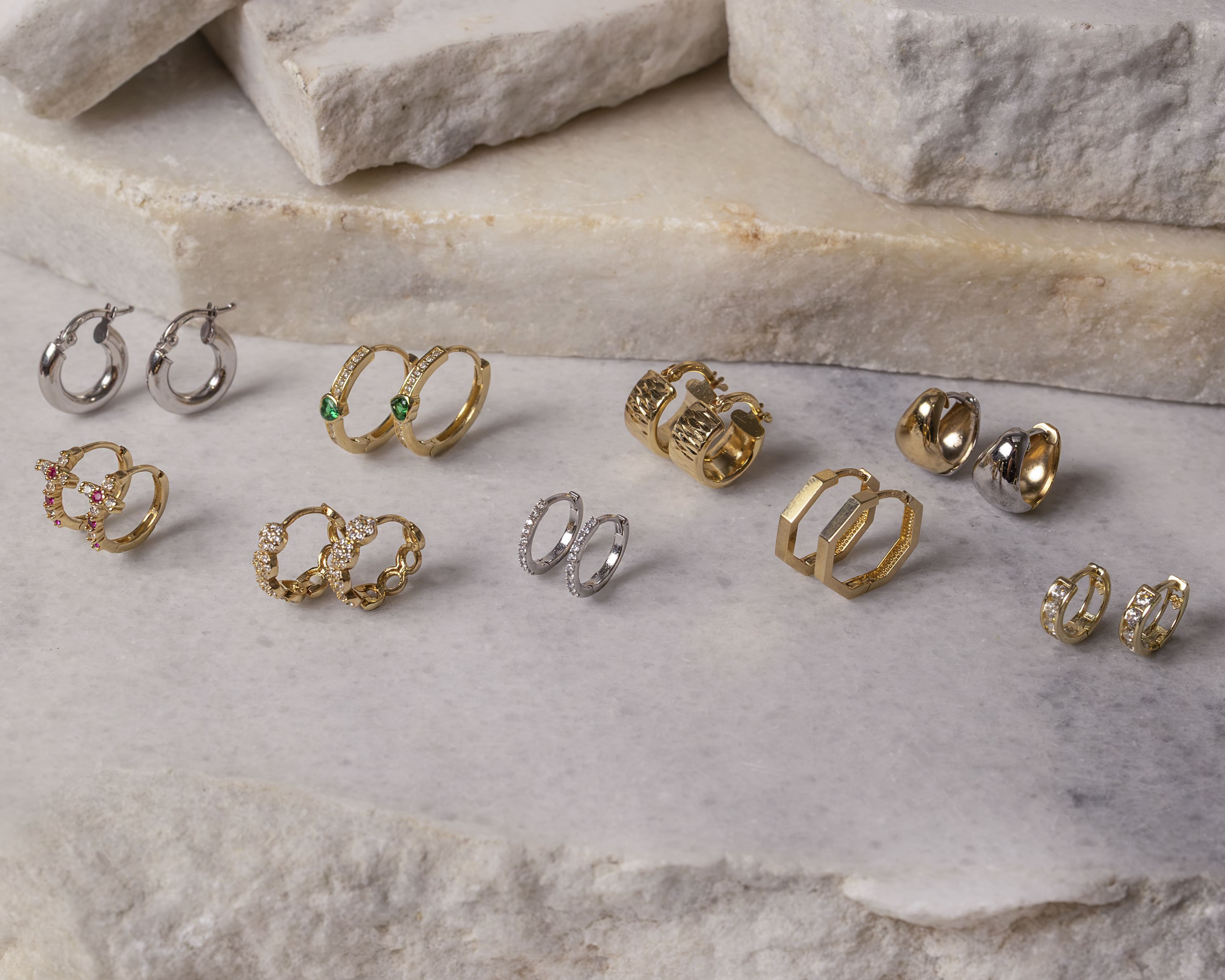 Eleven pairs of gold and silver hoop earrings with various designs and gemstones displayed on a white marble surface with stacked stone slabs in the background.