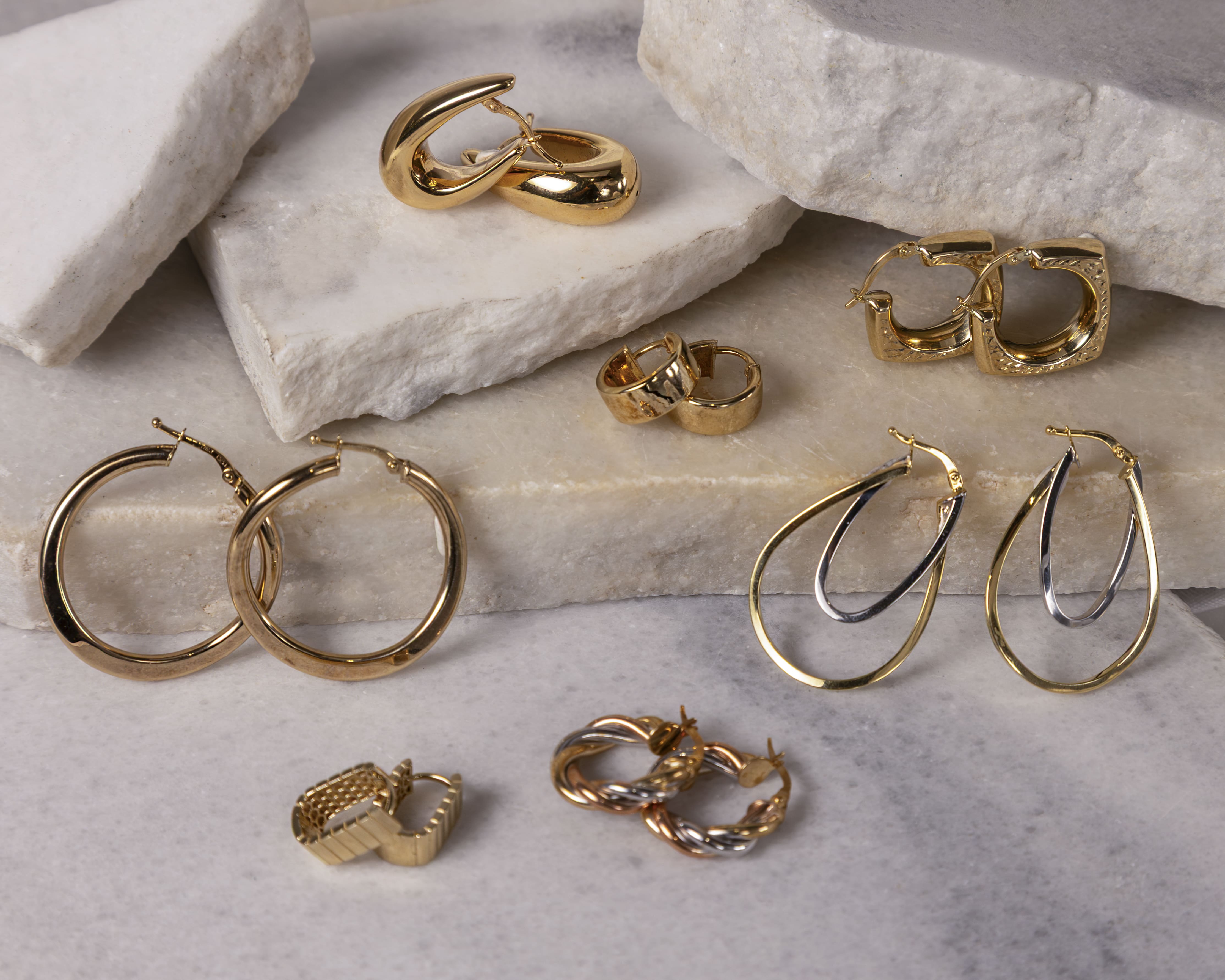 Various pairs of gold hoop earrings arranged on white stone slabs including smooth, twisted, mixed metal, and textured designs.