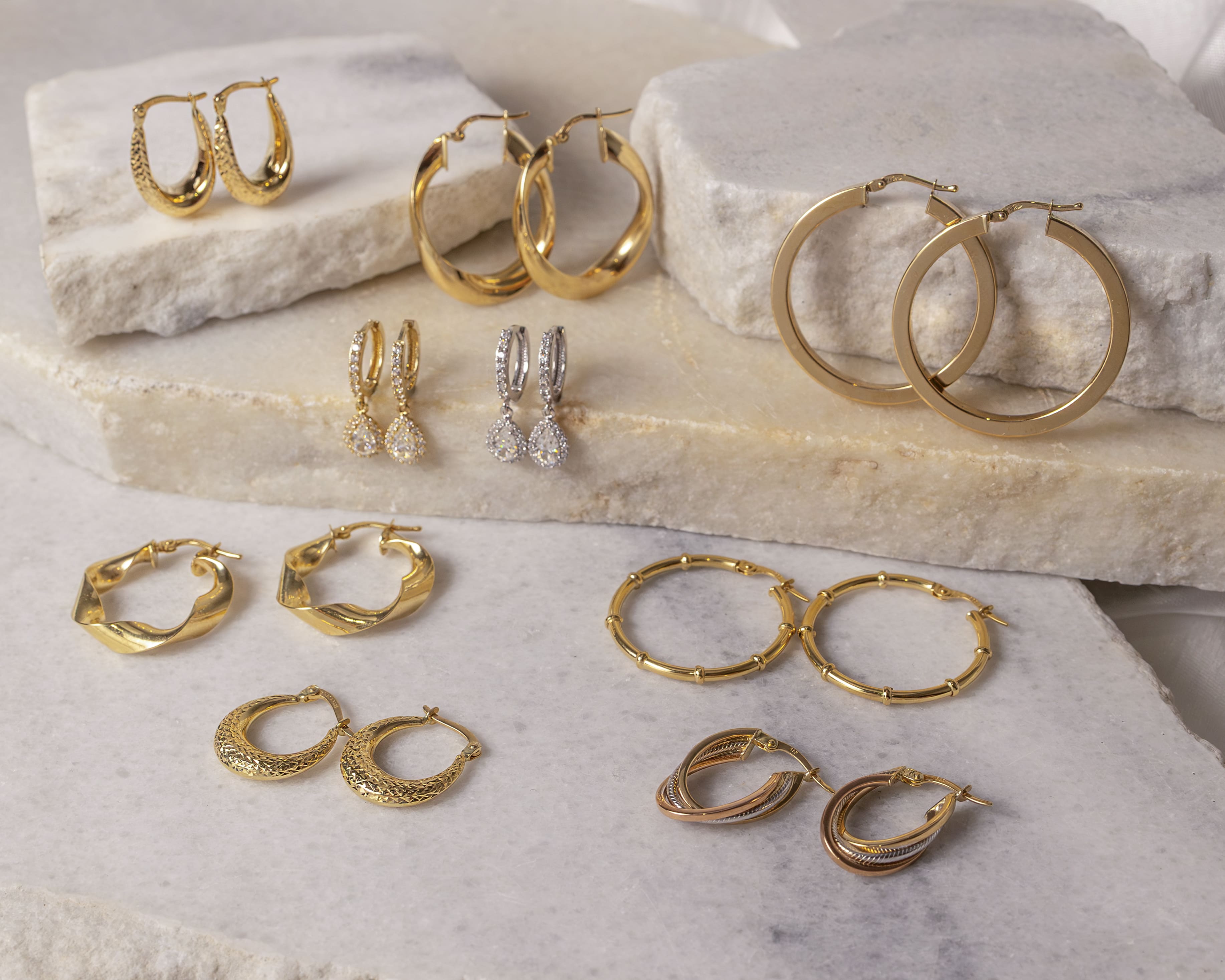 Various pairs of gold hoop earrings including textured, twisted, plain, and some with dangling teardrop-shaped crystals displayed on layered stone slabs.