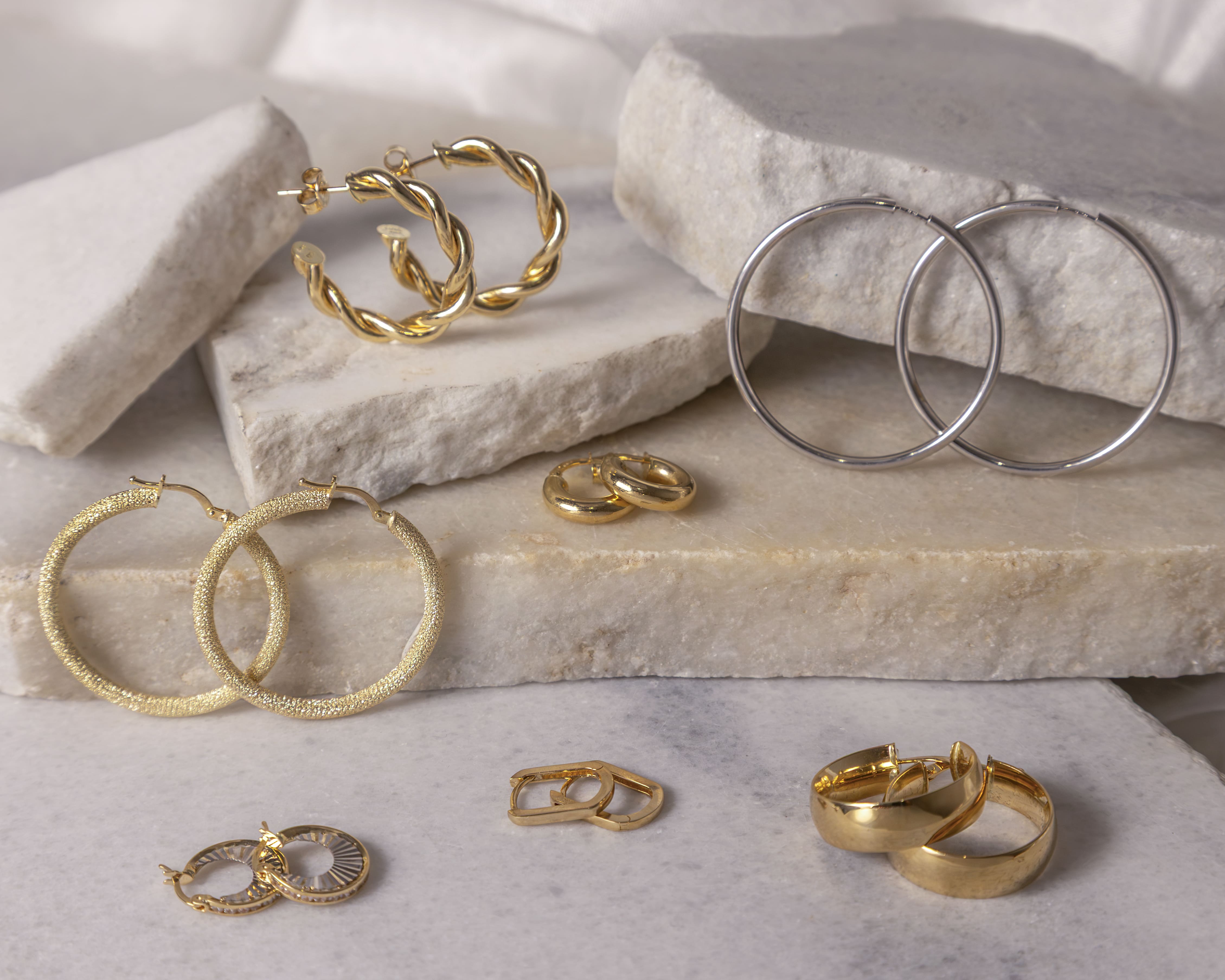 Seven pairs of gold and silver hoop earrings arranged on white stone slabs with various textures and sizes.