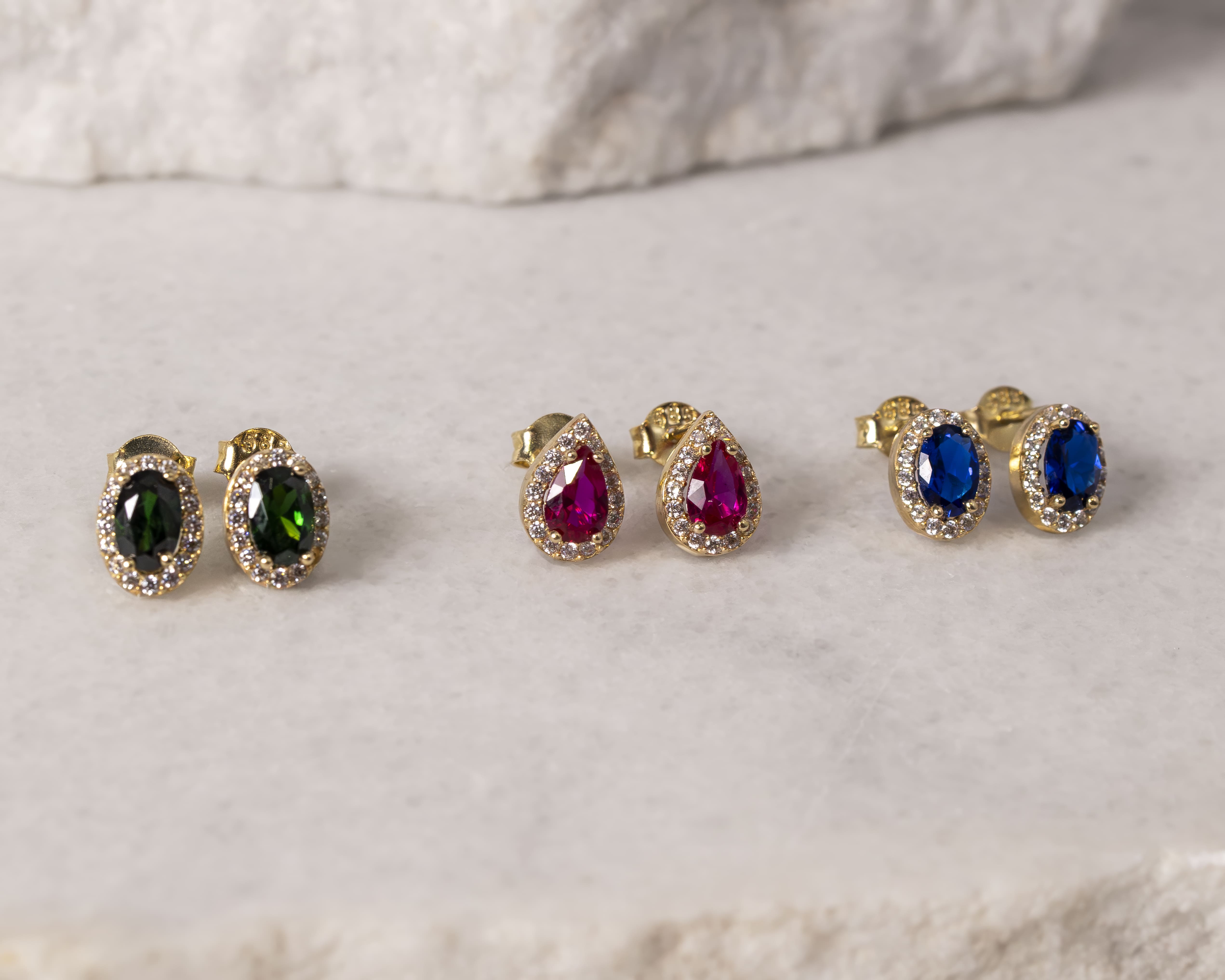 Three pairs of gold stud earrings with oval gemstones in green, teardrop-shaped red, and oval blue surrounded by small clear stones, on a light stone surface.