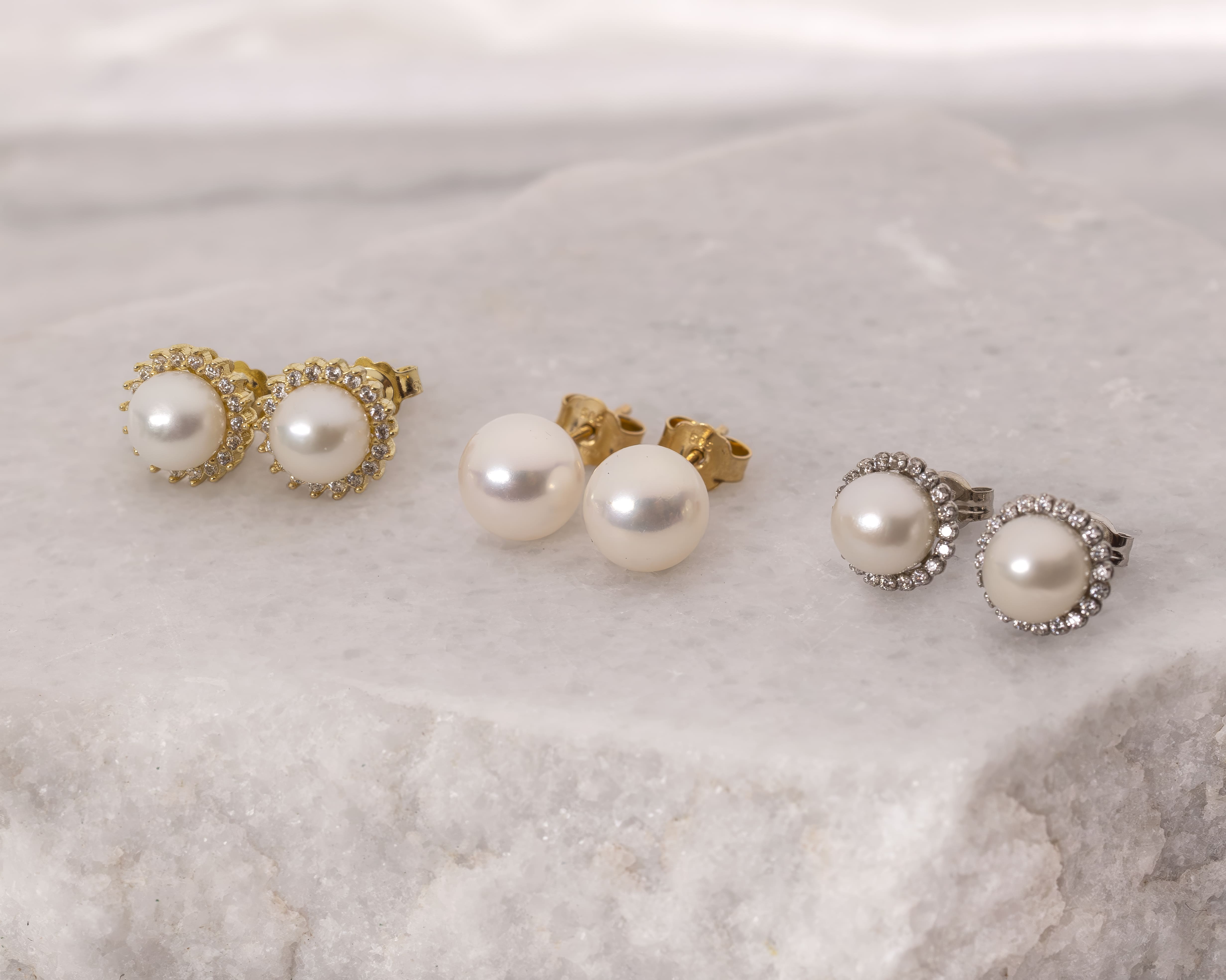 Three pairs of pearl stud earrings on a light marble surface, two pairs surrounded by small gemstones in gold and silver settings.