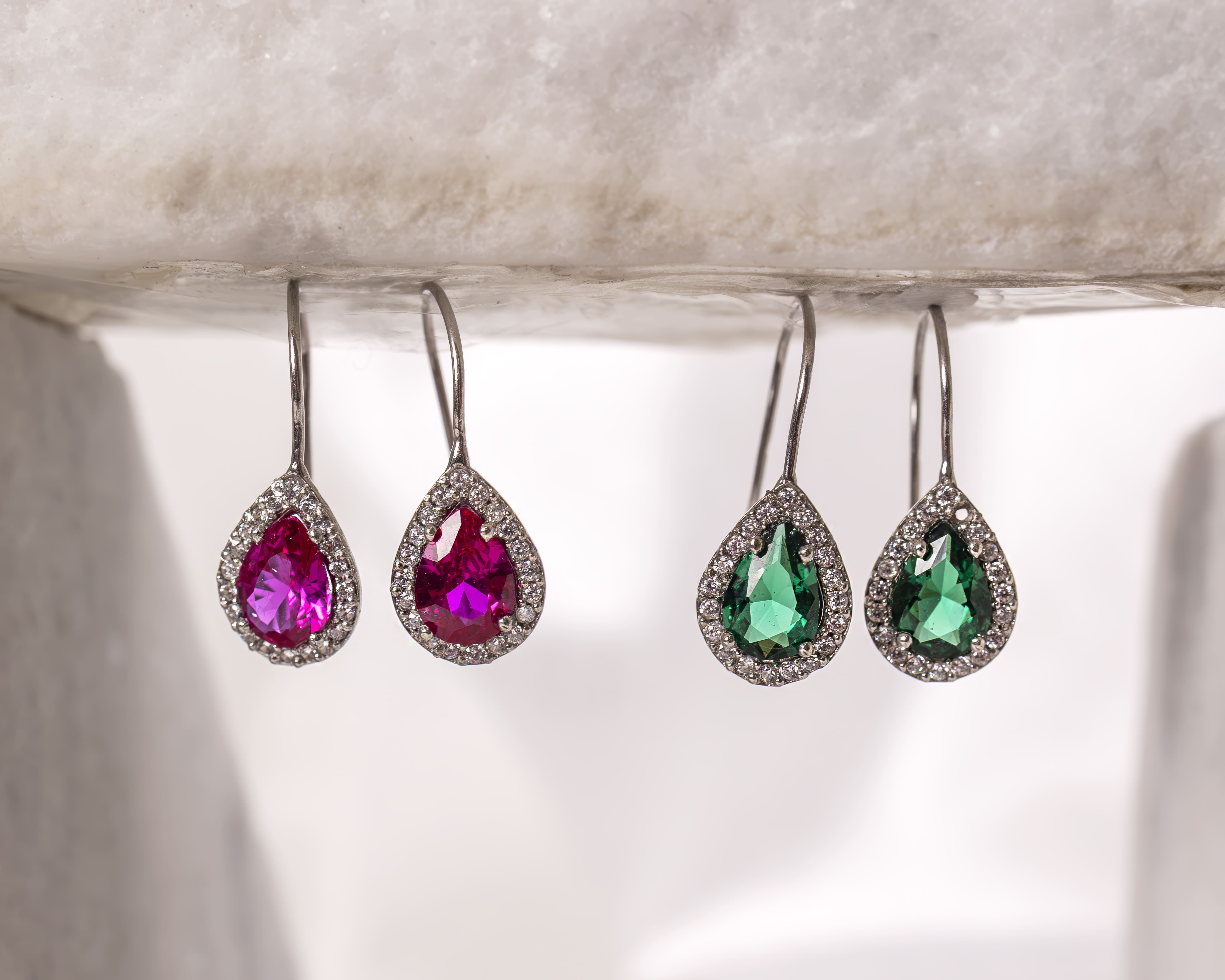 Two pairs of teardrop-shaped earrings with gem centers, one pair with pink stones and the other with green stones, both surrounded by small clear crystals.