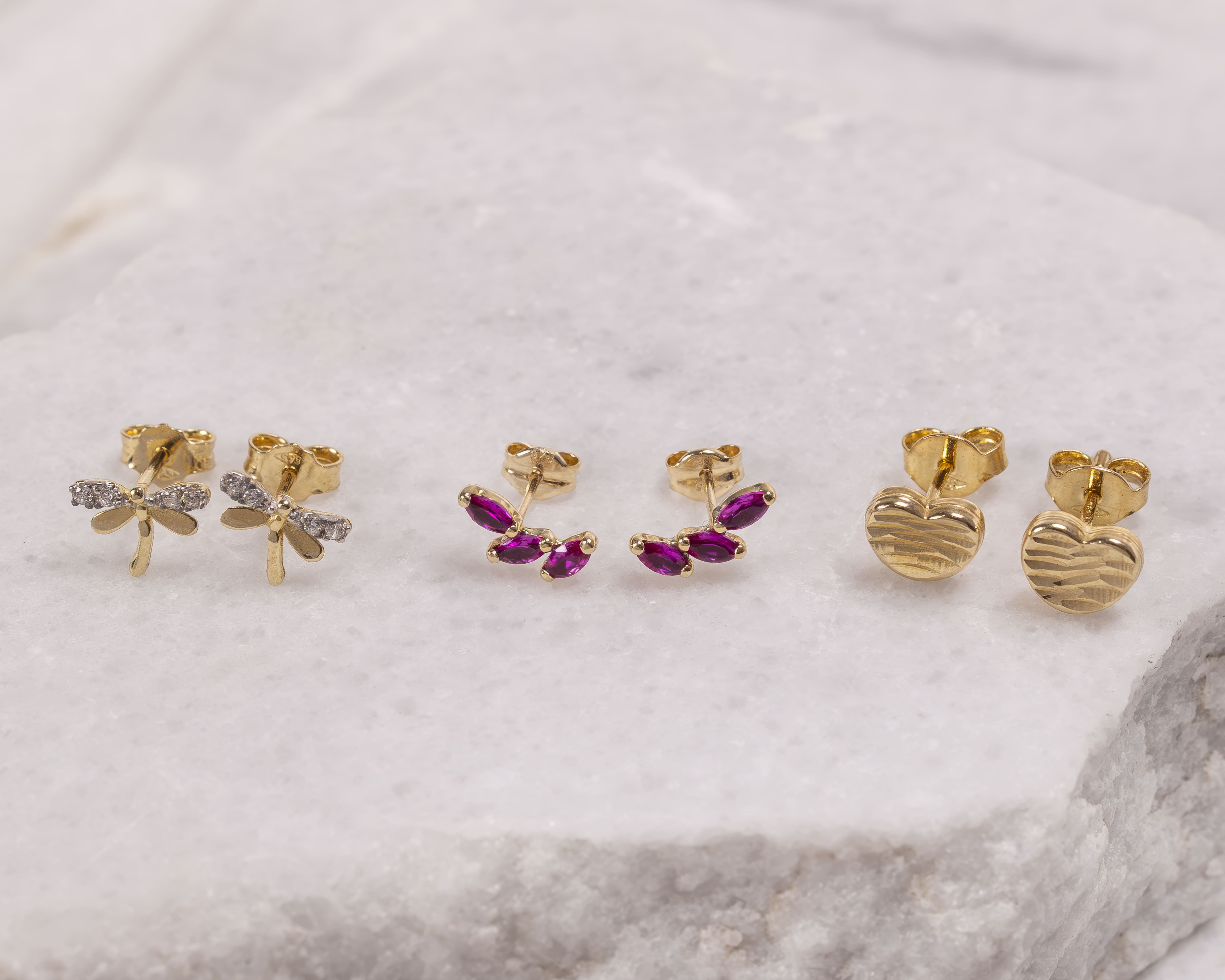 Three pairs of gold stud earrings on white marble: dragonflies with diamond wings, purple gemstone clusters, and textured hearts.