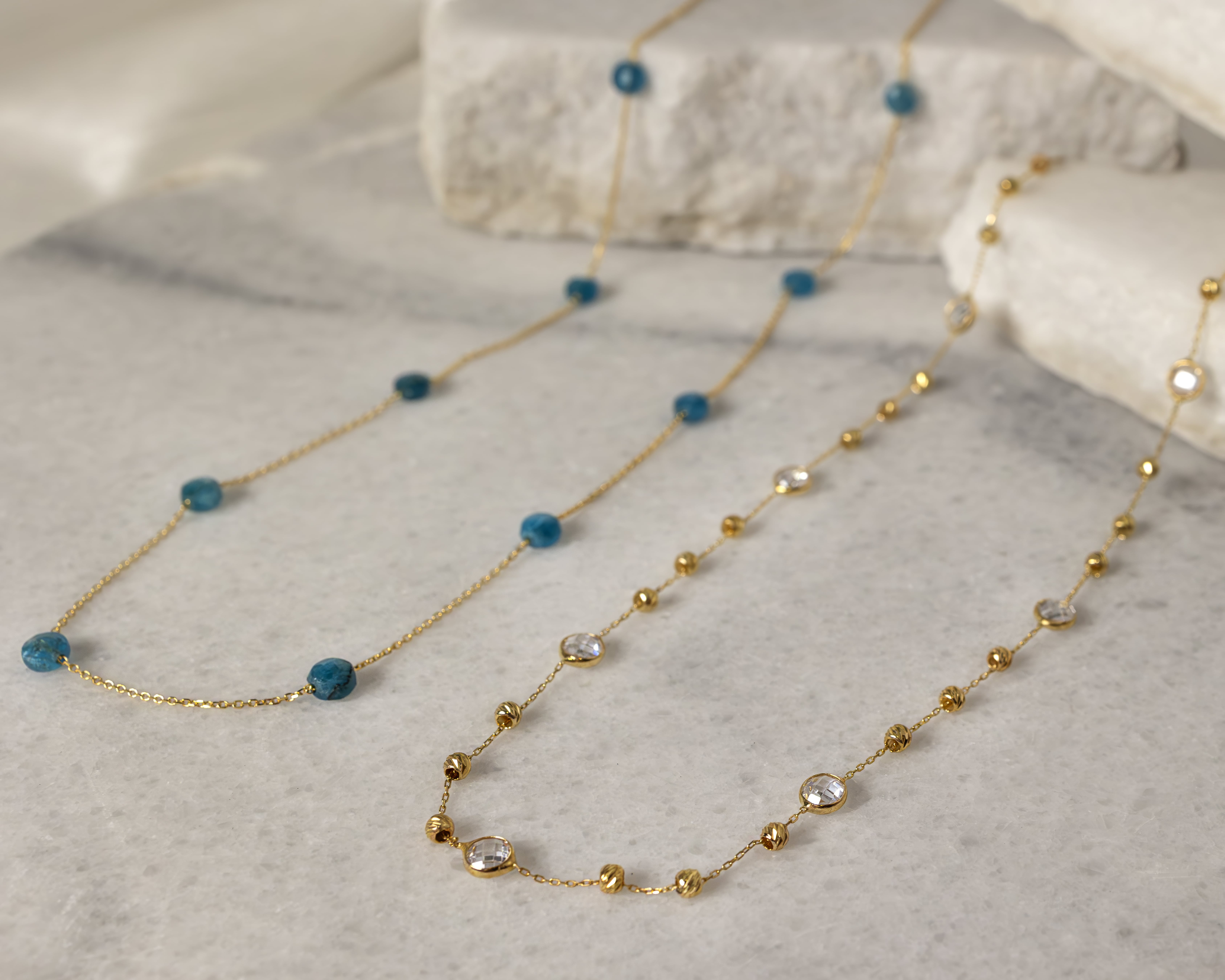Two delicate gold chain necklaces on a light marble surface, one with blue beads and the other with clear gem and gold bead accents.