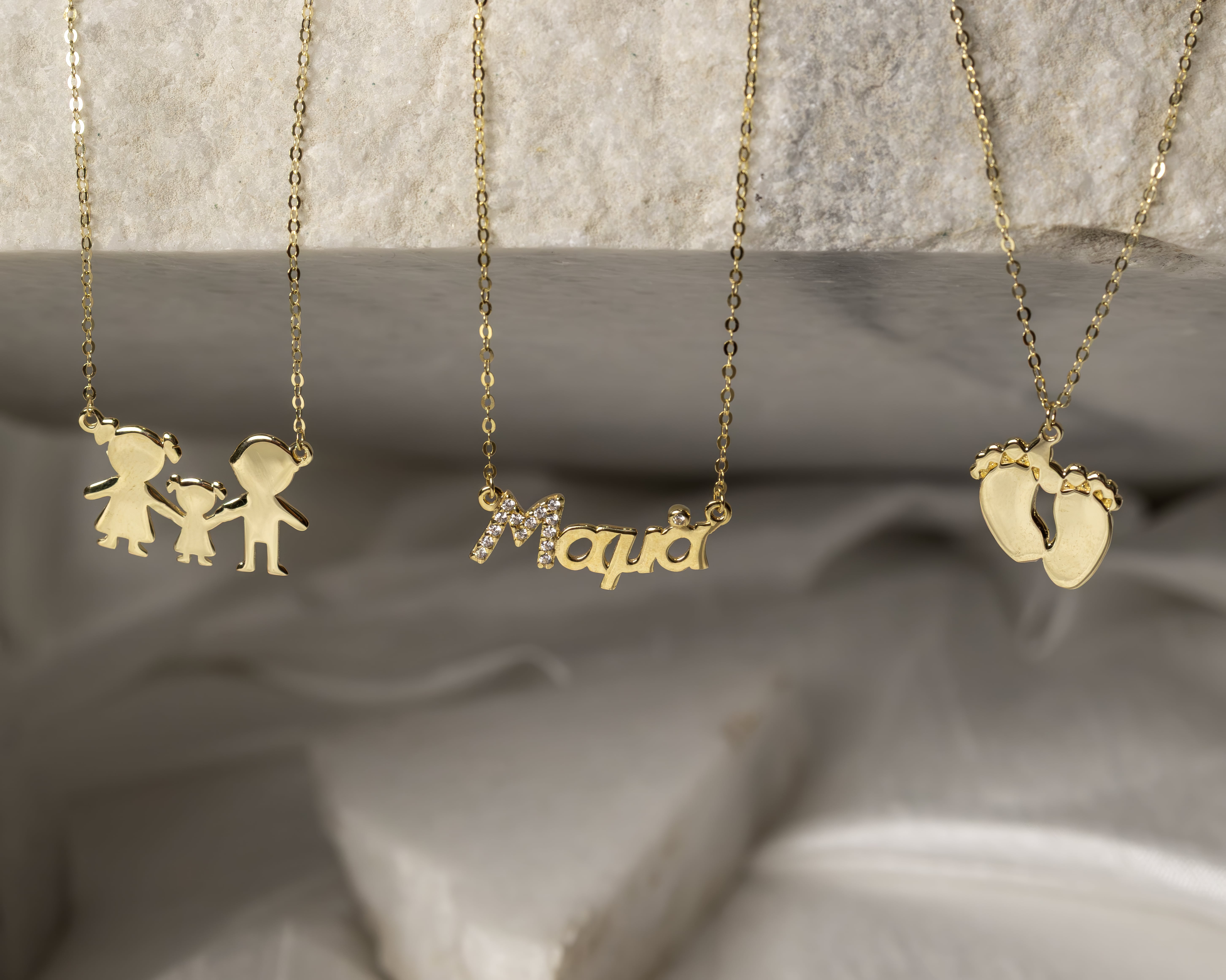 Three gold necklaces hanging, one with family figure charms, one with the word 'Mama' in script with small diamonds, and one with baby feet charms.