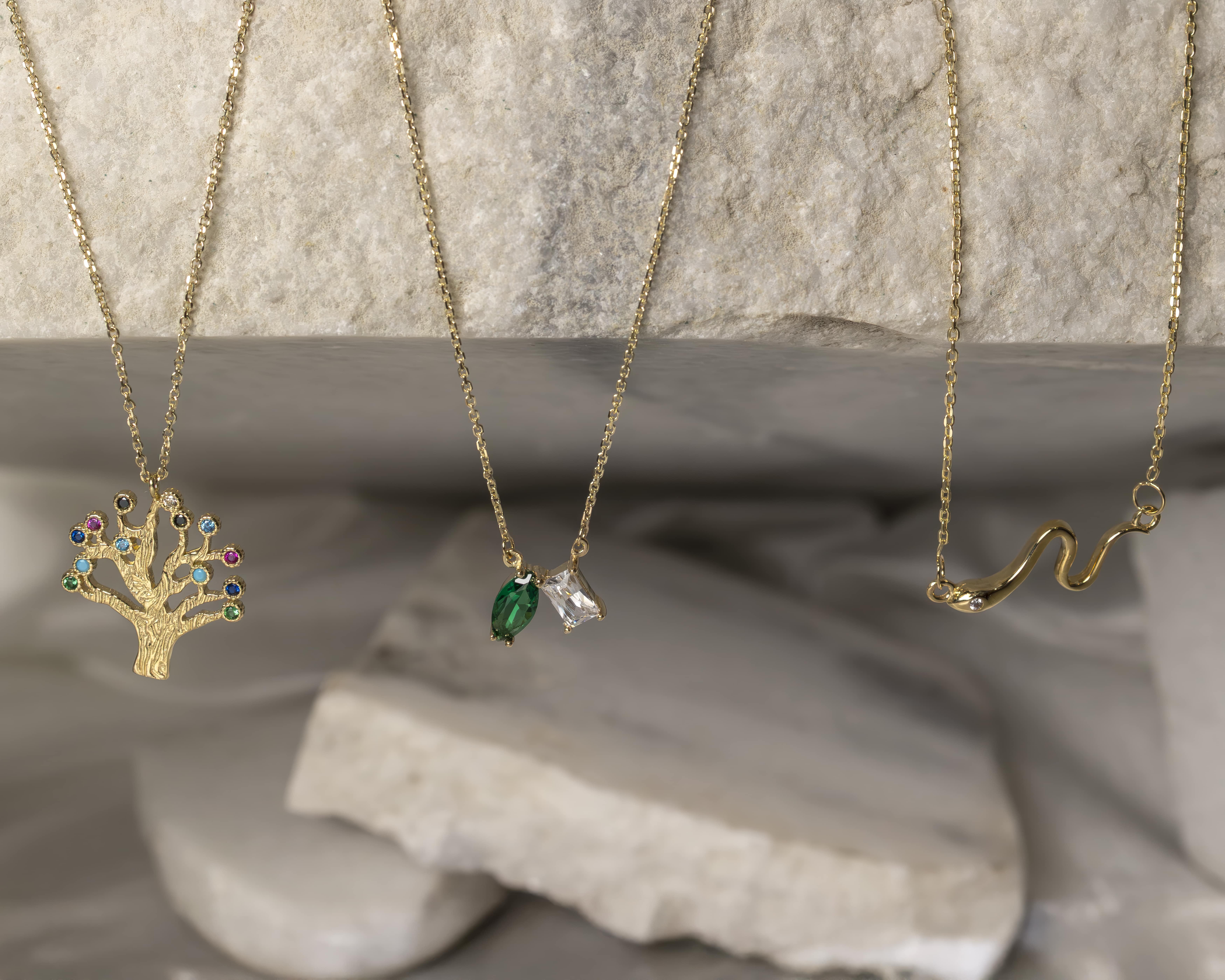 Three gold necklaces hanging against a stone background: left with a tree pendant adorned with multicolored gems, center with two marquise-shaped green and clear stones, and right with a snake-shaped pendant.