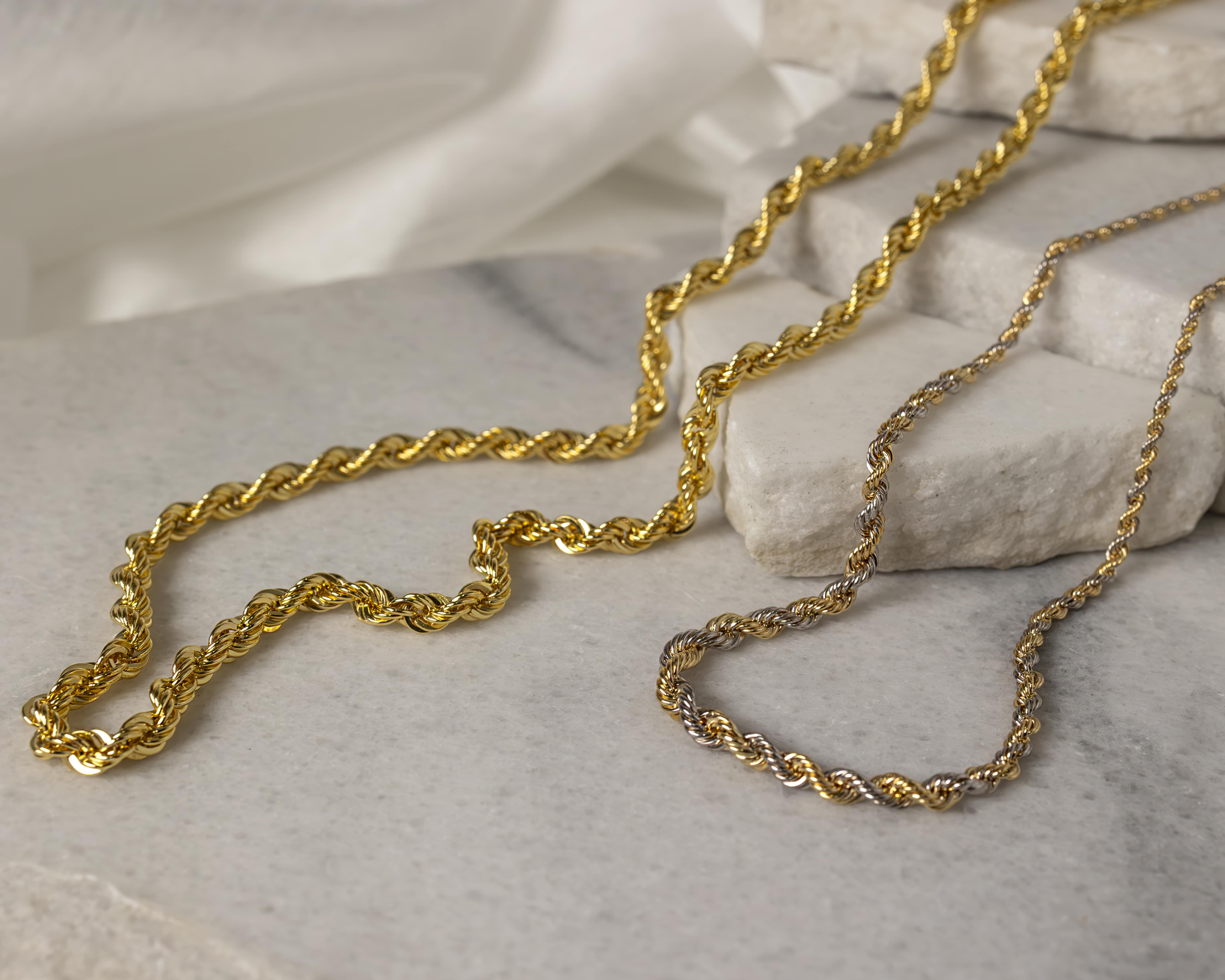 Two twisted rope chains, one solid gold and one gold with silver accents, displayed on a light stone surface.