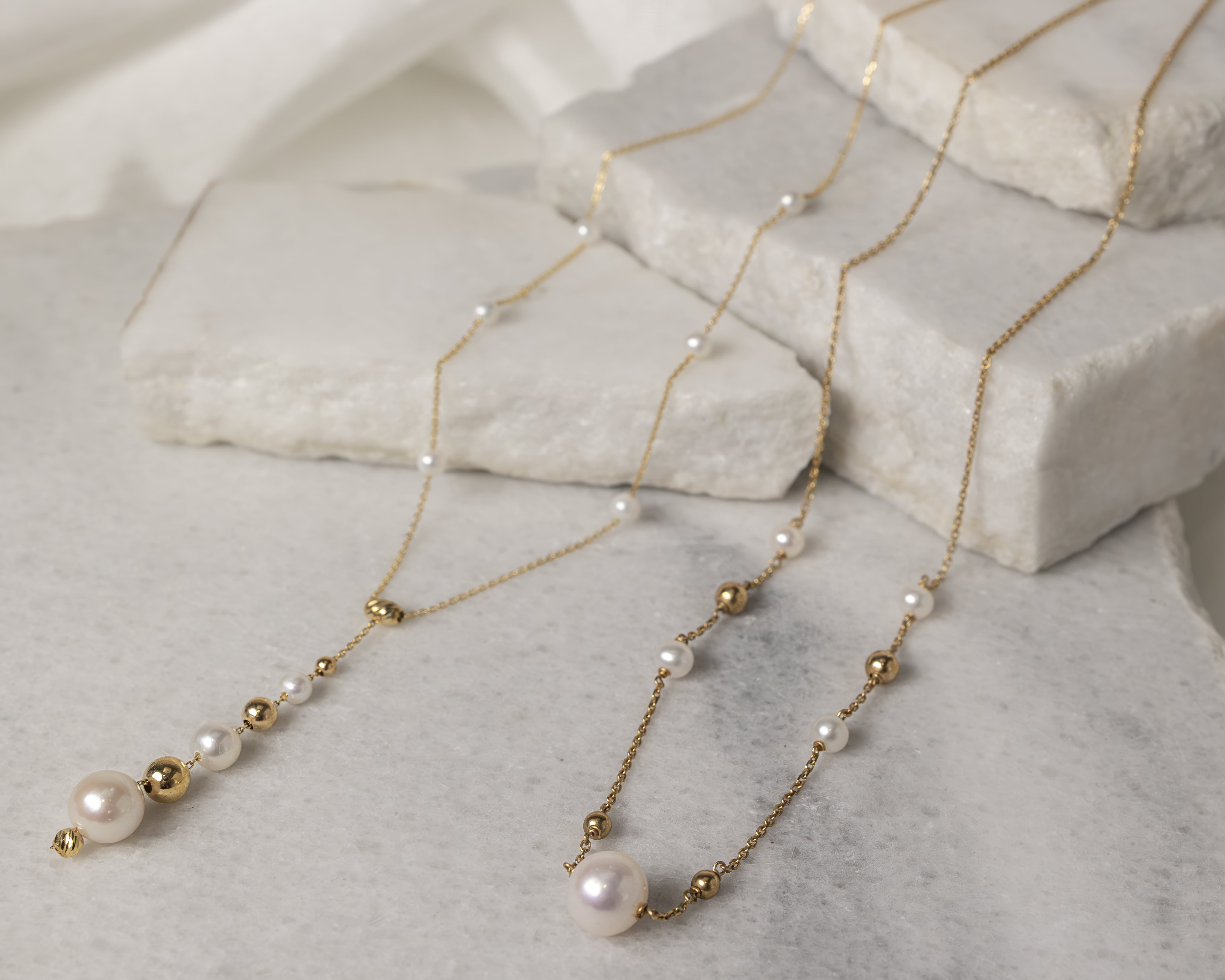 Two delicate gold necklaces with alternating small white pearls and gold beads displayed on white marble slabs.