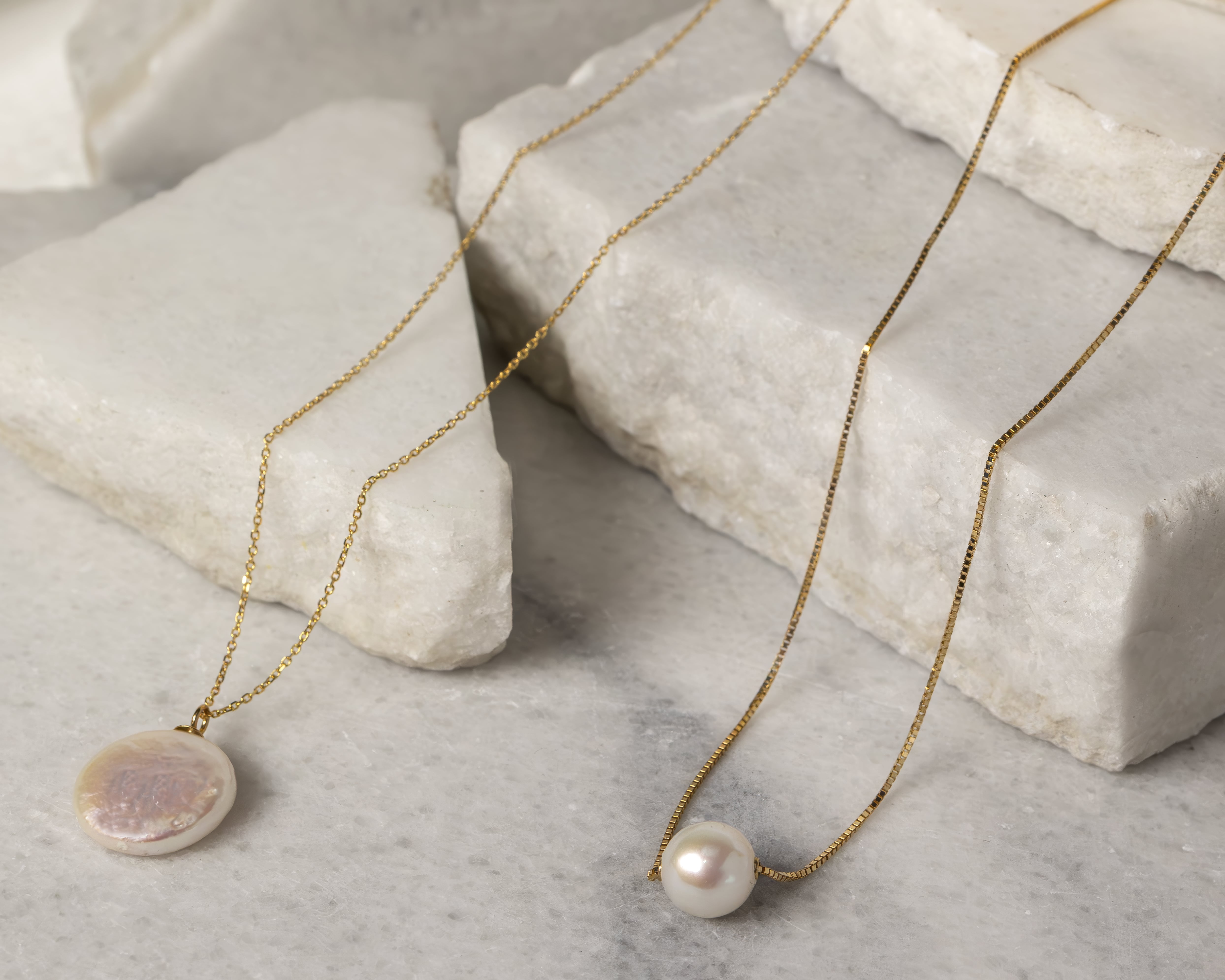 Two gold chain necklaces each with a pearl pendant displayed on white marble slabs.
