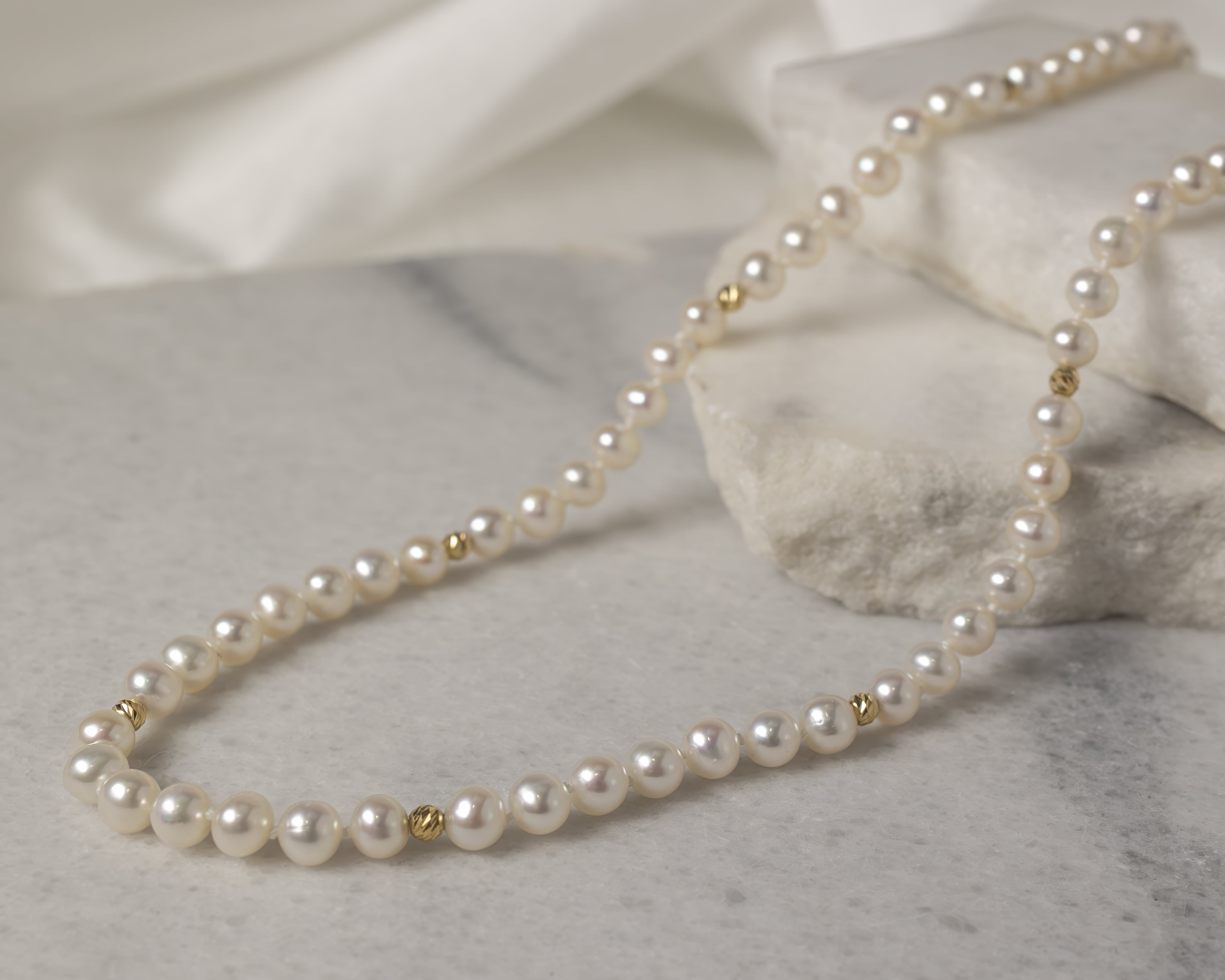 Elegant pearl necklace with small gold beads placed on a marble surface with stacked stone slabs in the background.