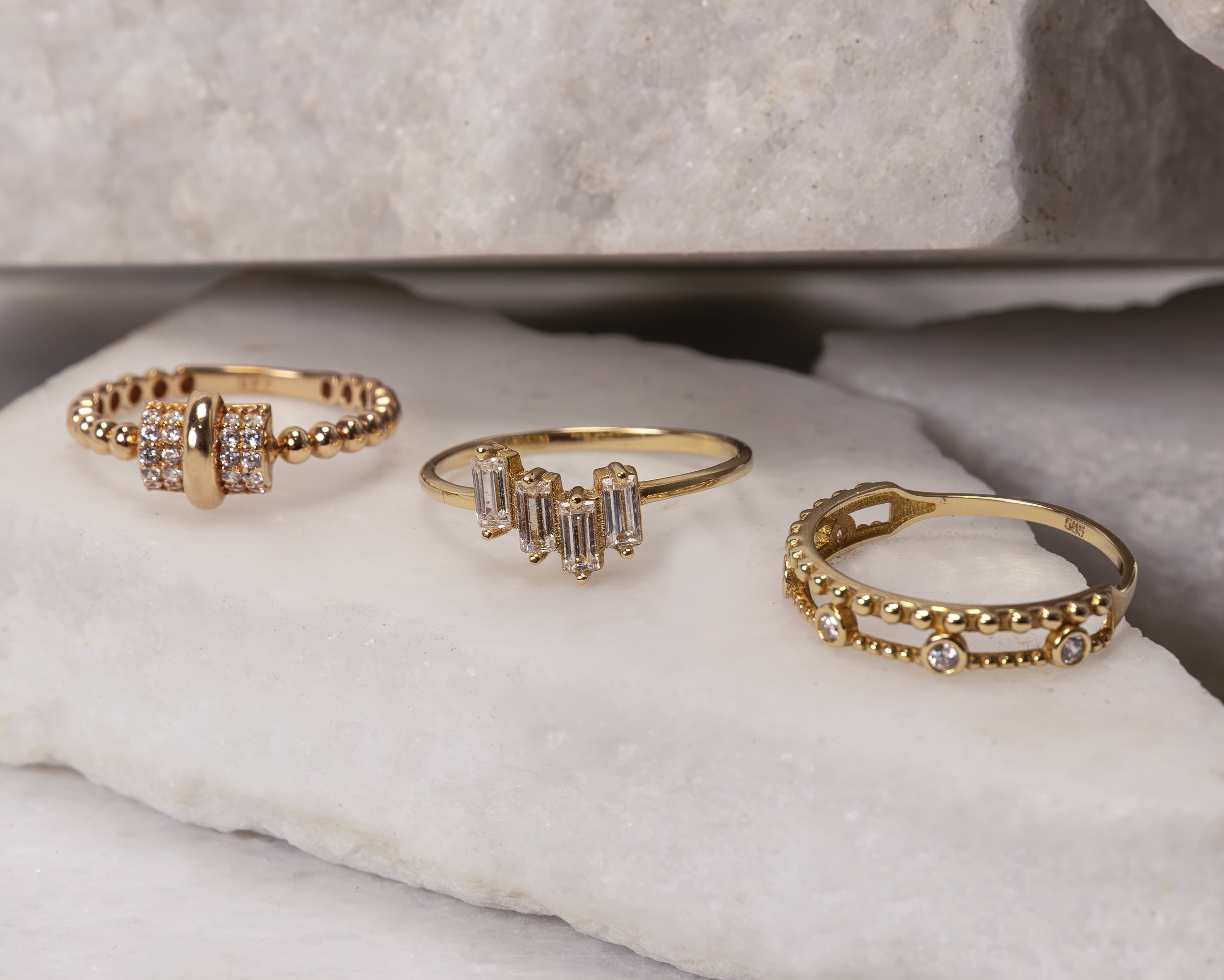 Three gold rings with diamond accents displayed on white marble slabs.