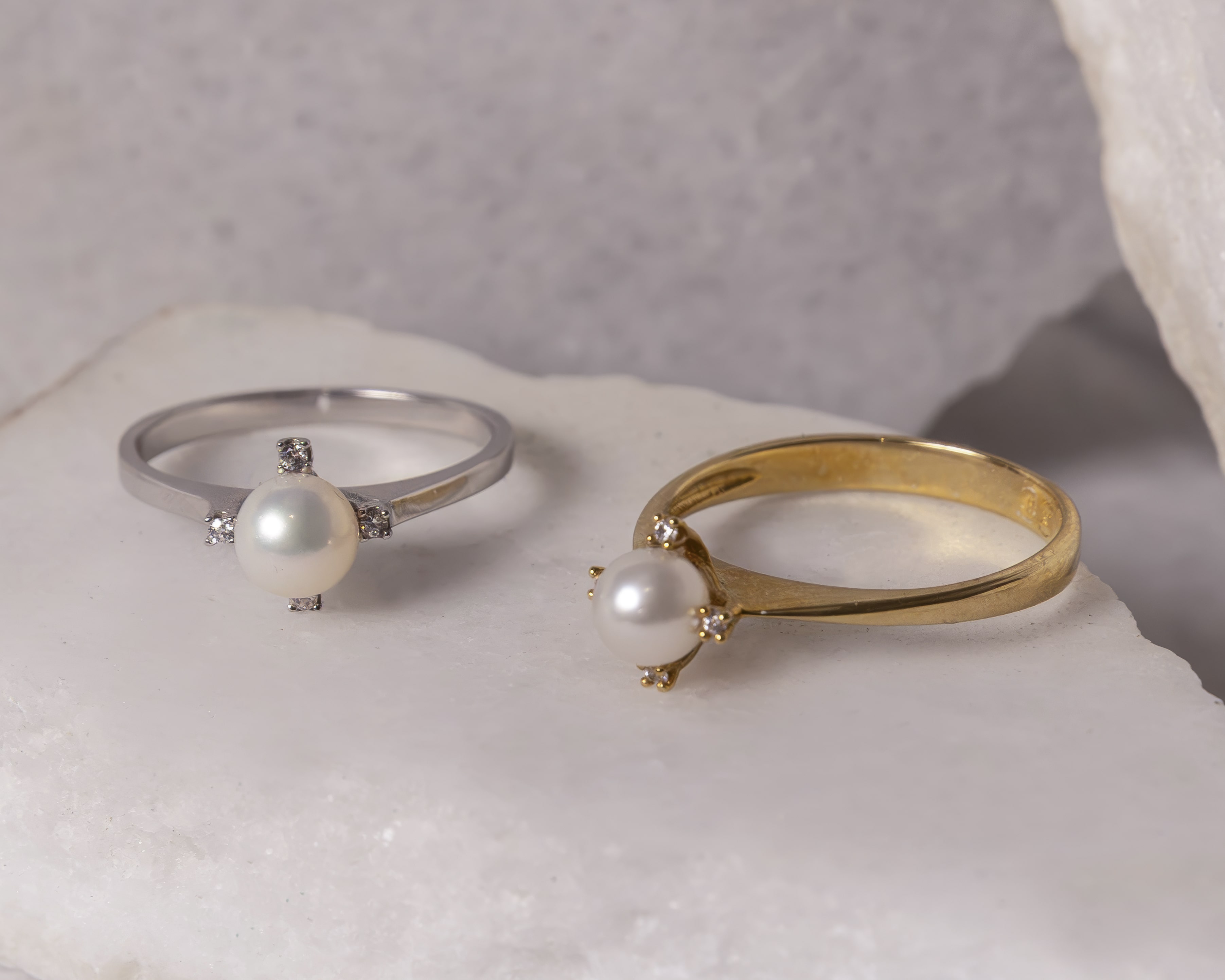 Two pearl rings on a white stone surface, one with a silver band and the other with a gold band, both adorned with small diamonds.