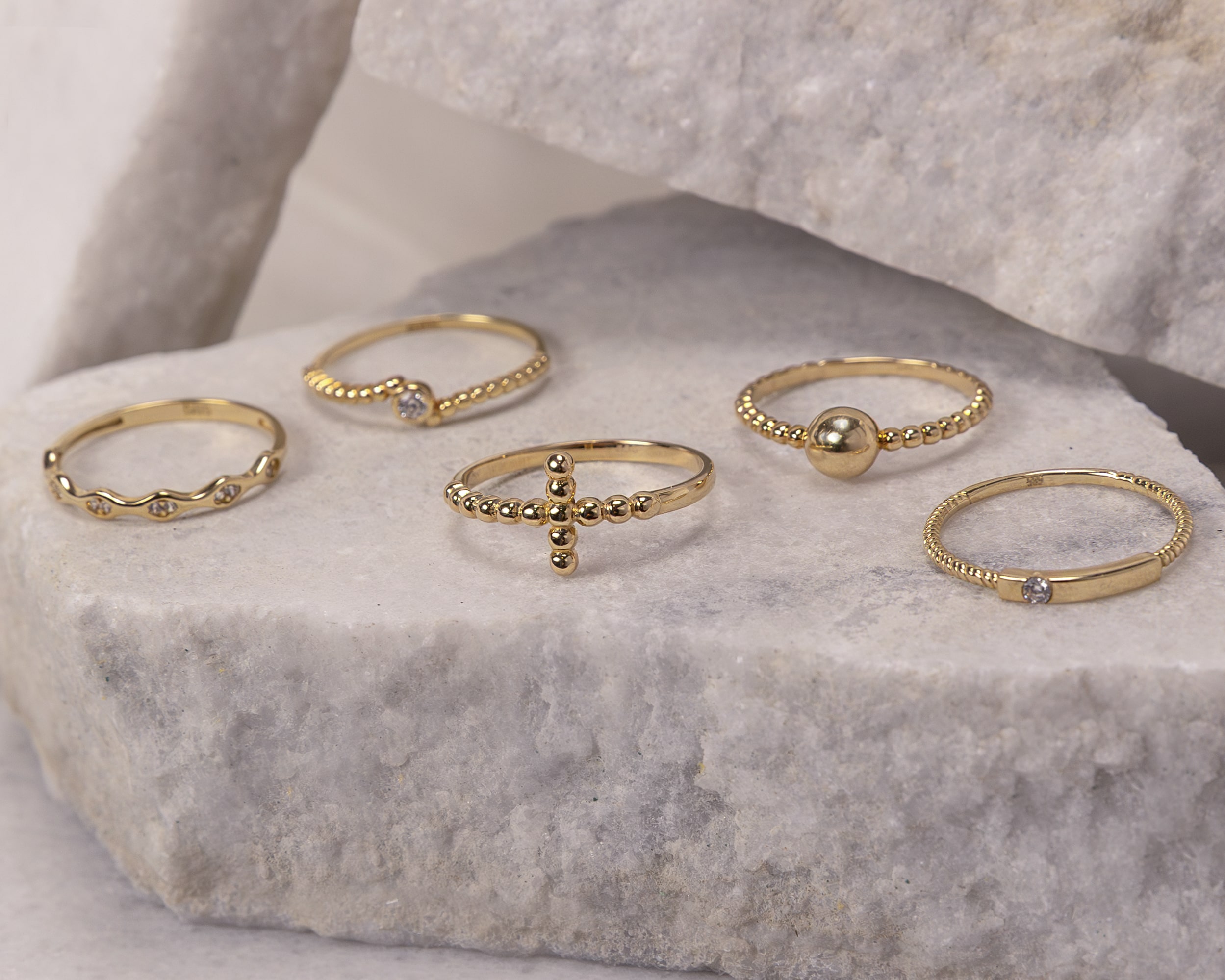 Five delicate gold rings with beaded and textured designs displayed on light gray stone surfaces.